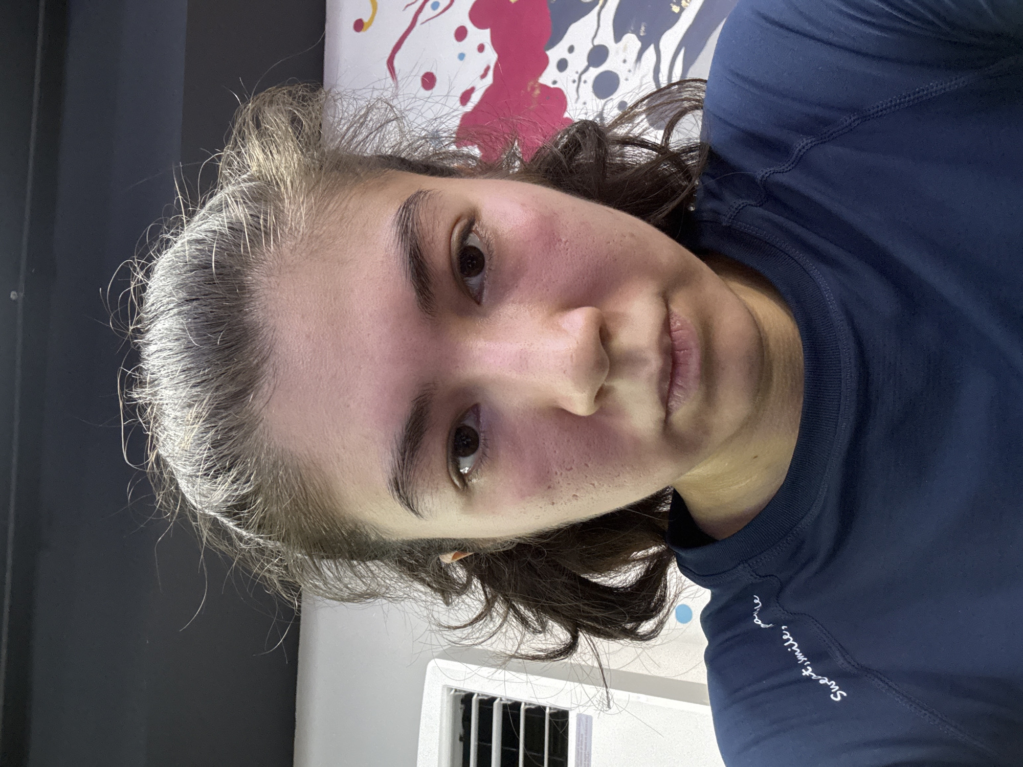 A young woman is captured in a close-up, self-taken photograph. She has brown hair pulled back loosely, with some strands framing her face. Her eyes are dark brown, and she has prominent eyebrows. Her face appears flushed, particularly on her cheeks and around her nose. She is wearing a dark blue long-sleeved shirt with white text on the left shoulder that reads "Sweat, smile, pour." In the background, a white air conditioning unit is visible on the left, and a colorful abstract mural with pink, white, and blue splatters is on the right. The lighting is somewhat harsh, casting shadows. The location is unknown, and the subject appears to be in an indoor setting, possibly a gym or a studio.