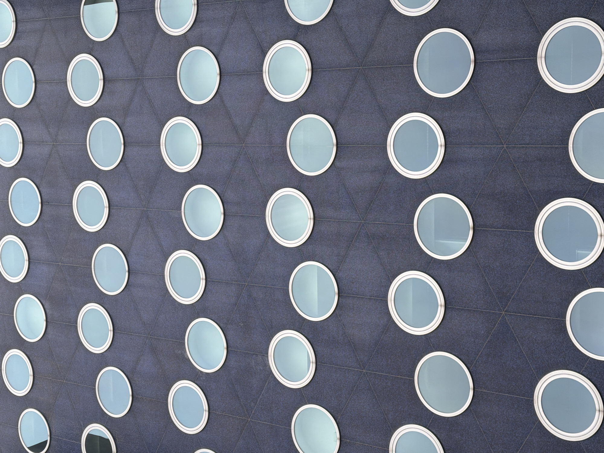 The image shows a close-up, low-angle view of a dark blue, textured facade of a building, likely an office building or apartment complex, featuring a repeating pattern of circular windows. The windows are evenly spaced in a grid-like fashion, with a slight offset between rows, creating a tessellated effect. Each window has a white, metallic frame and is filled with a light blue glass, reflecting the sky. The overall impression is one of modern architectural design.
While the provided image is a general architectural detail, this specific facade design is characteristic of the former Rabobank building located at Van der Kooijplein 1, 2287 DE Rijswijk, Netherlands. This building, designed by architects Mecanoo, was completed in 1995 and is known for its distinctive circular windows, which create a playful and organic appearance against the dark facade. The building is situated in the Plaspoelpolder business district of Rijswijk. There are no people or specific actions visible in this image, only the static architectural elements of the building's exterior.