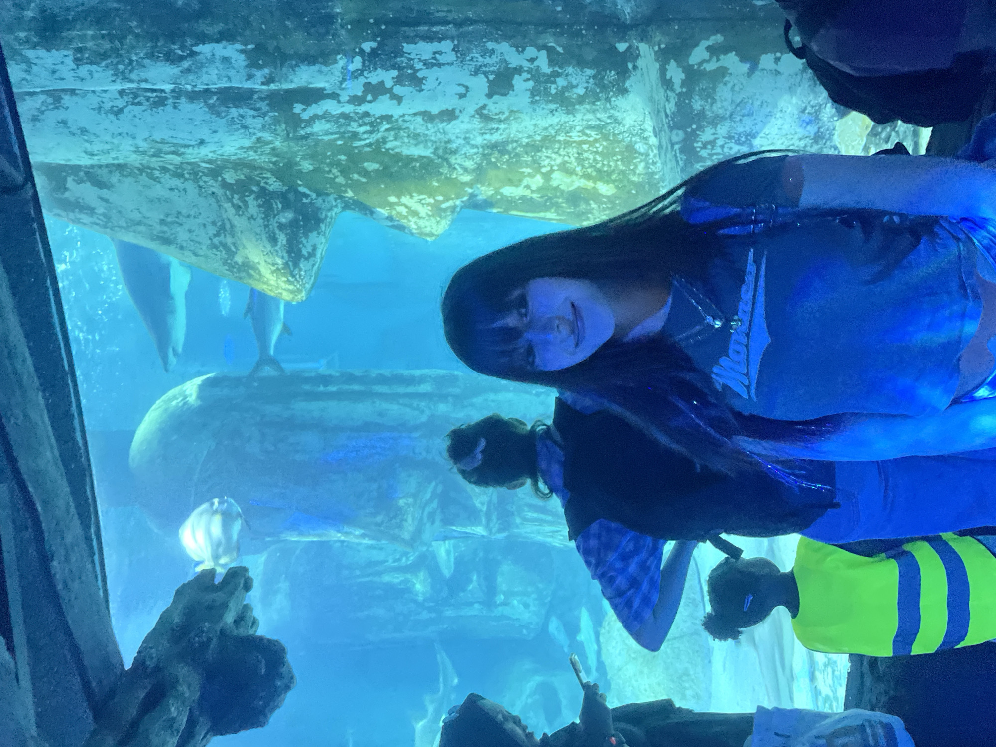 An aquarium scene is depicted, with a young woman in the foreground looking towards the camera. She has long, dark hair with bangs and is wearing a dark t-shirt with a white logo and a necklace. Behind her, other individuals are visible, including a child wearing a bright yellow vest with blue stripes and a person in a blue and black checkered shirt. The environment is an underwater exhibit, with large rock formations and various fish swimming in the blue-tinted water. The lighting is dim and atmospheric. The location is identified as the Sea Life London Aquarium, located on the South Bank of the River Thames in London, United Kingdom.