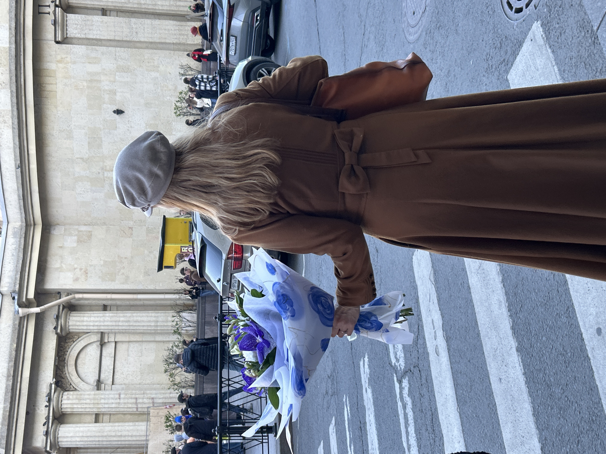 The image, rotated vertically, captures a street scene in Saint Petersburg, Russia, focusing on a woman seen from behind. She is wearing a long, warm brown coat with a bow detail at the back of the waist, and a light grey beret. Her long, wavy blonde hair flows from beneath the beret. She carries a brown leather shoulder bag and holds a bouquet of flowers wrapped in decorative white paper with blue floral patterns. The flowers themselves appear to be purple and white.
The woman is positioned near a pedestrian crosswalk with prominent white stripes. In the background, a large, ornate building with classical columns and a stone facade stands tall, suggesting historical architecture common in Saint Petersburg. Several other people are visible on the sidewalk and near the building entrance. Parked cars are also in the background; a license plate "707РТ 178" and "Х901АА 178" are visible on two different vehicles, with "178" being the regional code for Saint Petersburg, Russia. The scene appears to be taking place during the daytime, under soft lighting, indicating an overcast or perhaps late afternoon sky, with no harsh shadows. No specific event is clearly depicted beyond general street activity.