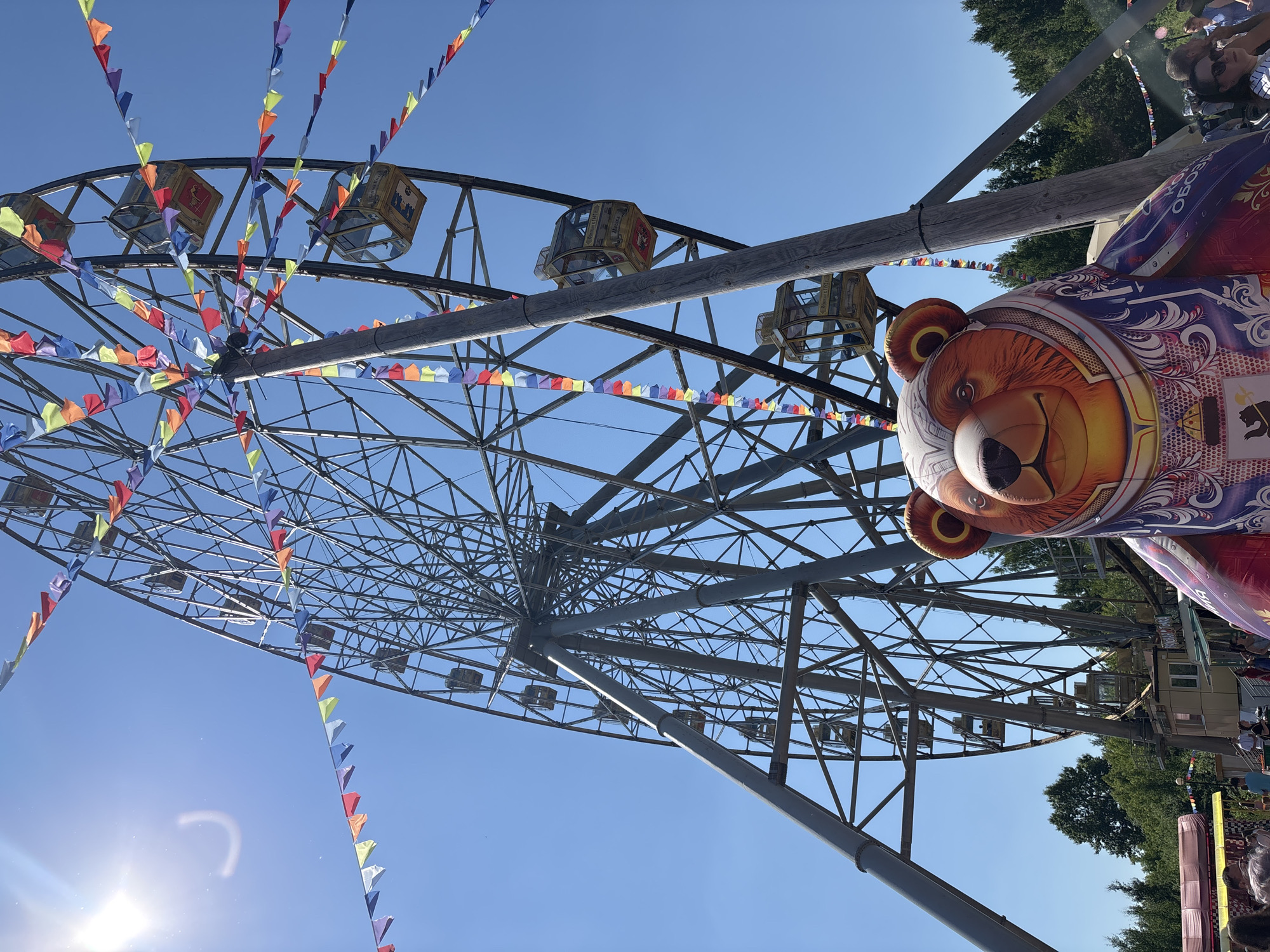 An outdoor, low-angle, full shot captures a ferris wheel dominating the scene under a clear blue sky. Numerous colorful triangular flags are strung across the frame, attached to the ferris wheel's structure and poles. The ferris wheel is a large, metallic structure with many spokes and supports, featuring several enclosed yellow cabins. In the foreground, a large, inflatable sculpture of a bear's head and upper torso is positioned slightly to the right of center, with its face looking forward. The bear is decorated with intricate patterns. Below the ferris wheel, there's a glimpse of a park or fairground with trees lining the background and some structures and people visible at the lower edge. The sun is prominently featured in the upper left quadrant of the image, casting a bright glare. The location is identified as Rusanovo, Russia.