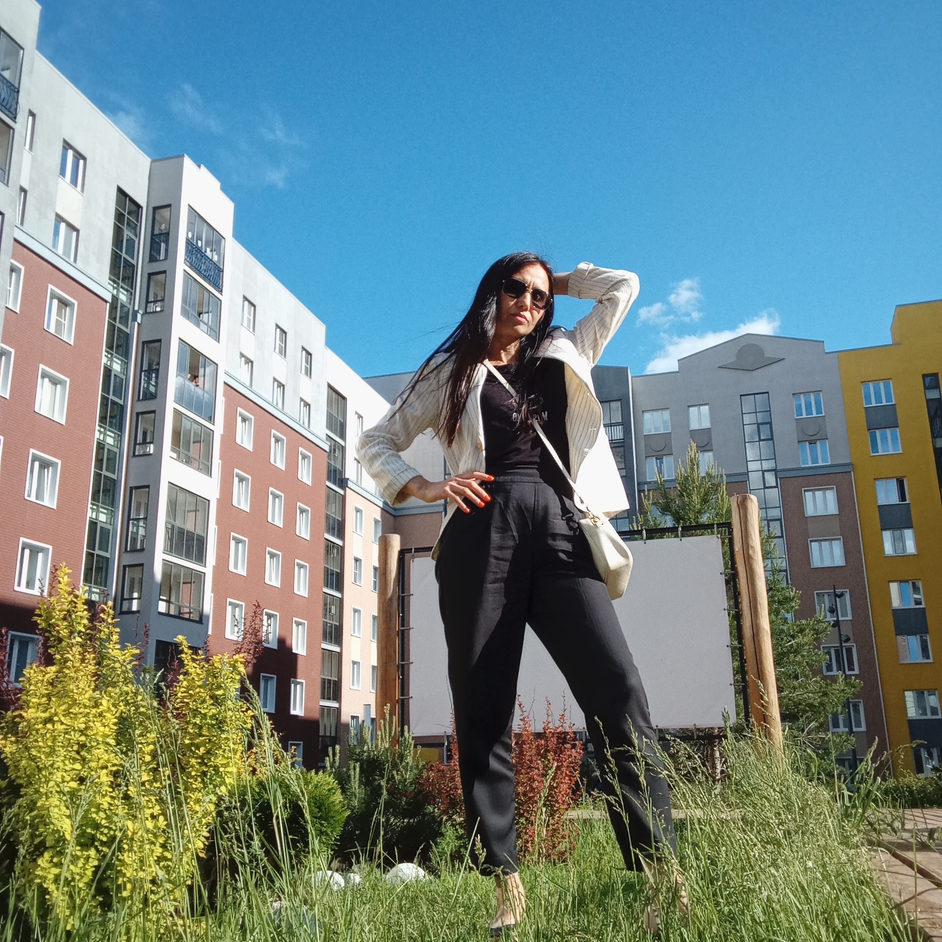 An outdoor, eye-level shot captures a woman posing in a grassy area with modern residential buildings in the background under a bright blue sky, likely in Pervomaysky, Russia. The woman, positioned centrally, wears a black top, black trousers, and a white striped blazer, complemented by black sunglasses and a light-colored crossbody bag. Her left hand rests on her hip, while her right hand is raised to her head, adjusting her long, dark hair. Her fingernails are painted red.
The setting appears to be a contemporary urban residential complex, characterized by multi-story apartment buildings with varied facades: white, brick red, grey, and yellow sections. Large windows are prominent on all buildings. The foreground features vibrant green grass and yellow-leafed bushes, suggesting a well-maintained green space. Behind the woman, partially obscured by her, is a tall wooden pole next to a large, blank white sign or board.
The image conveys a sunny, clear day, likely midday or early afternoon given the bright lighting and the high position of the sun creating subtle shadows. The overall mood is casual and confident, with the woman's pose suggesting she is modeling for the camera. No discernible text is visible on any objects or buildings.