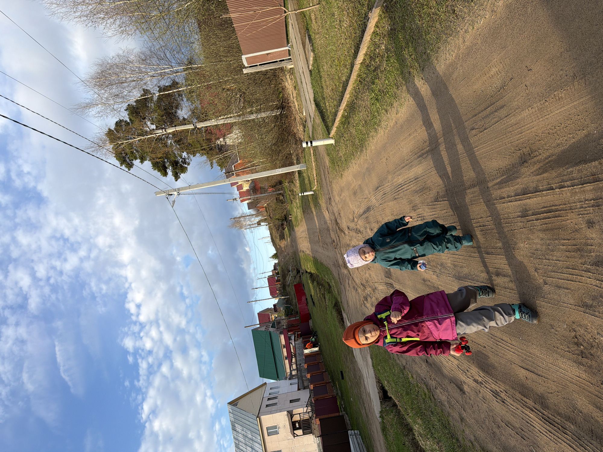 The image captures a rural or semi-rural scene in Chaikovsky, Russia, featuring two young children on a dirt road under a partly cloudy sky.
The main subjects are two children. One child, on the right, wears a dark green snowsuit and a patterned light-colored hat, holding a small blue or silver object. The other child, closer to the foreground and slightly to the left, is dressed in a magenta/purple jacket with bright yellow-green accents, grey pants, and an orange/brown hat, holding a red toy car. They both appear to be standing or walking on the unpaved road.
The scene unfolds on a wide dirt road showing distinct tire tracks, bordered by grassy verges on either side. Long shadows cast by the children stretch across the road, indicating a low sun, likely early morning or late afternoon. In the background, several houses and buildings of varying styles and colors are visible, some with distinctive green roofs. Utility poles with multiple wires line the road, and trees, some bare and some with green foliage, punctuate the landscape. The sky above is a mix of blue with scattered white clouds.
No visible text is present in the image. The overall impression is one of children enjoying an outdoor moment in a quiet village setting.