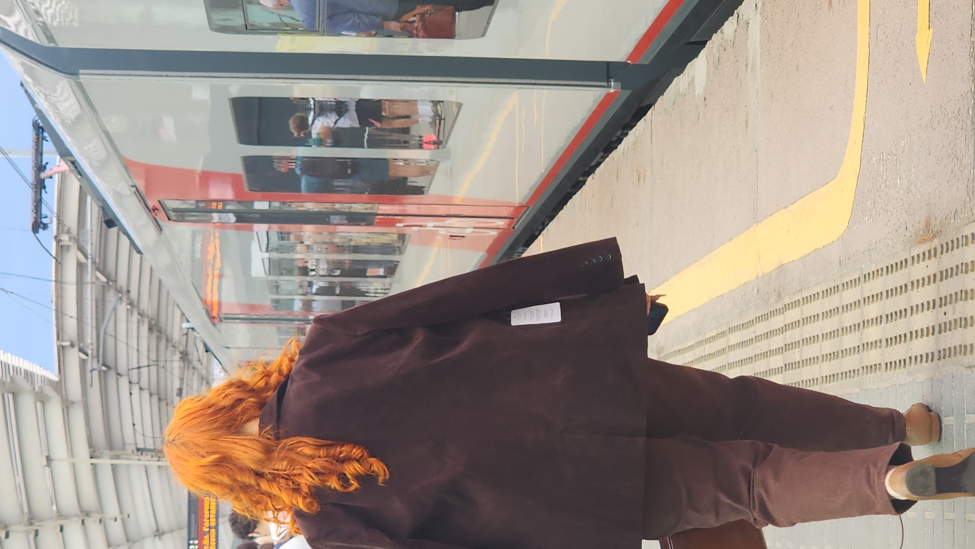 A person with bright orange curly hair walks away from the camera on a train platform in Moscow, Russia. The person is wearing a dark brown suit jacket and matching pants. The jacket has a tag hanging from the left sleeve. They are carrying a dark bag in their left hand and a dark phone in their right hand. The train is red and white and is stopped at the platform. Reflections of people can be seen in the train windows. The platform has a yellow line painted on it and textured tactile paving.