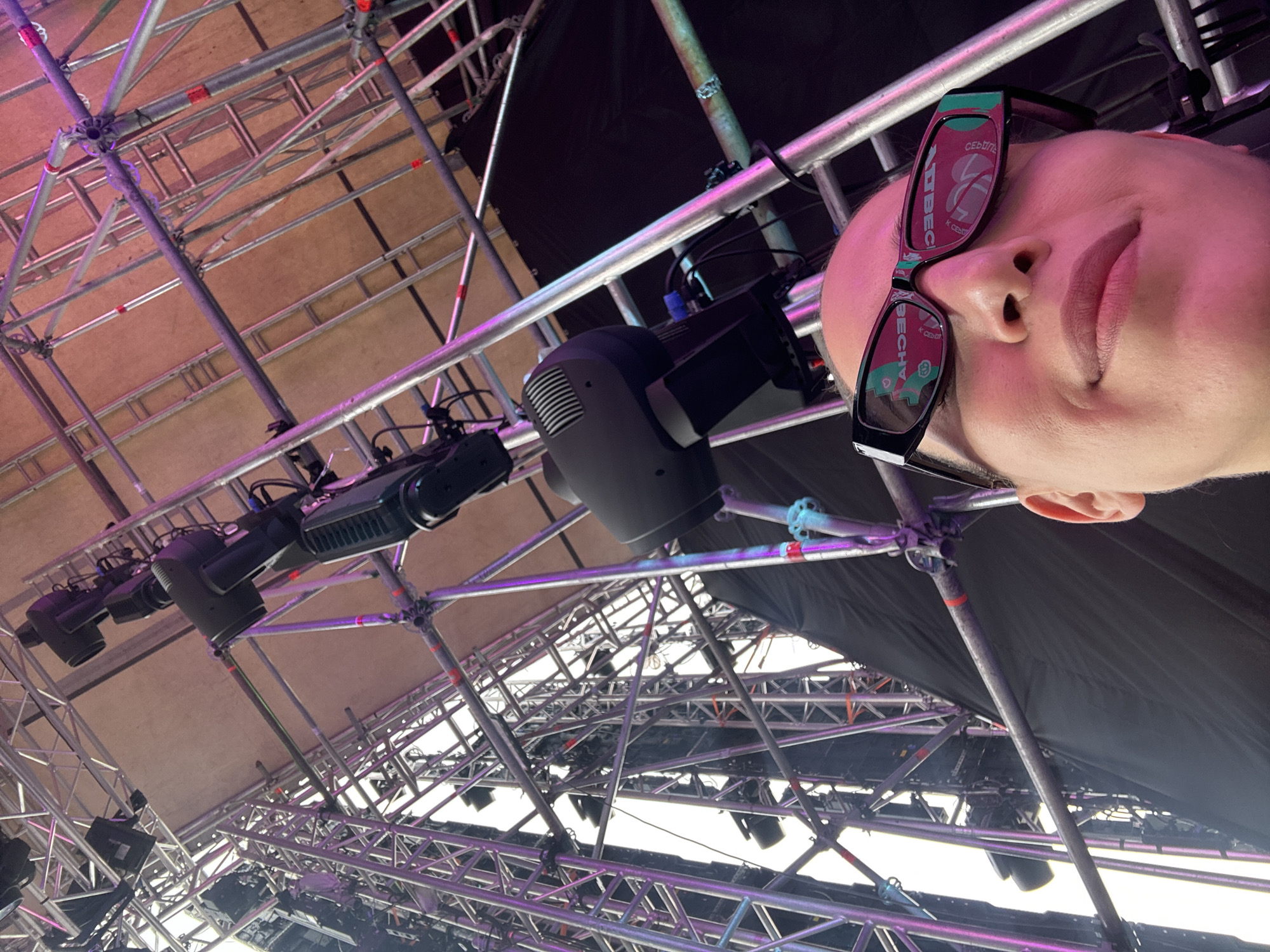 An outdoor, low-angle, selfie-style shot shows a person wearing black-rimmed sunglasses with reflective lenses, looking up towards a large, intricate scaffolding structure. The scaffolding is made of metal pipes and supports various lighting fixtures and what appear to be speakers or other stage equipment. The lights cast pink and purple hues on the structure. The background is a dark curtain or fabric. The person's face, upper lip, and nose are visible, and their expression is subtle, with a slight smile. The glasses reflect the pink and green colors of the surrounding stage lights. The scene appears to be a concert or event setup. There is no information to place this image in Khabarovsk, Russia.