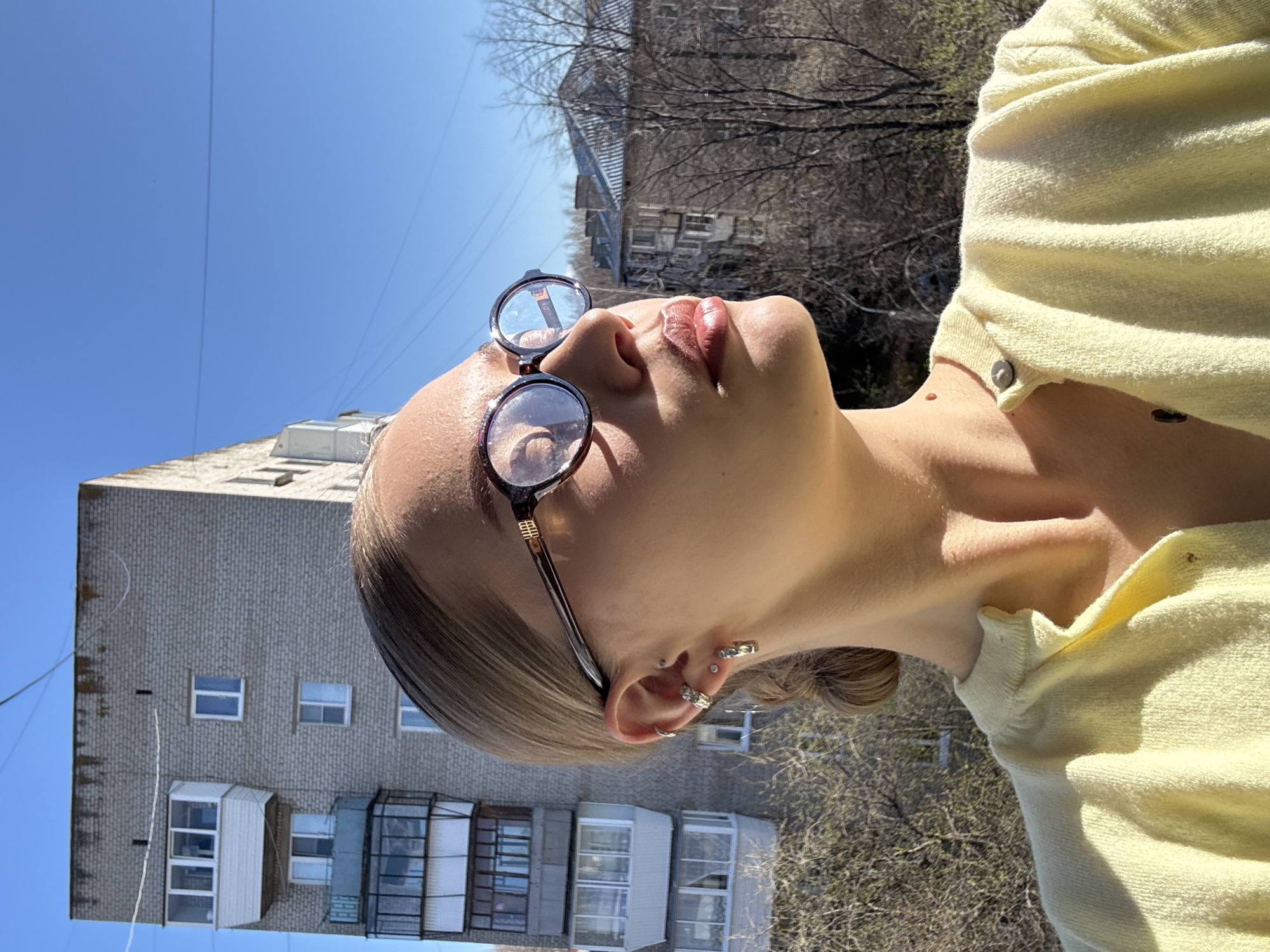 A woman with fair skin is wearing round glasses and a yellow cardigan, looking up at a clear blue sky. Her ear is adorned with multiple piercings. Behind her is a multi-story brick building with white framed windows and balconies. Bare trees are interspersed with the building and sky. The location is likely Chaikovsky, Russia.
