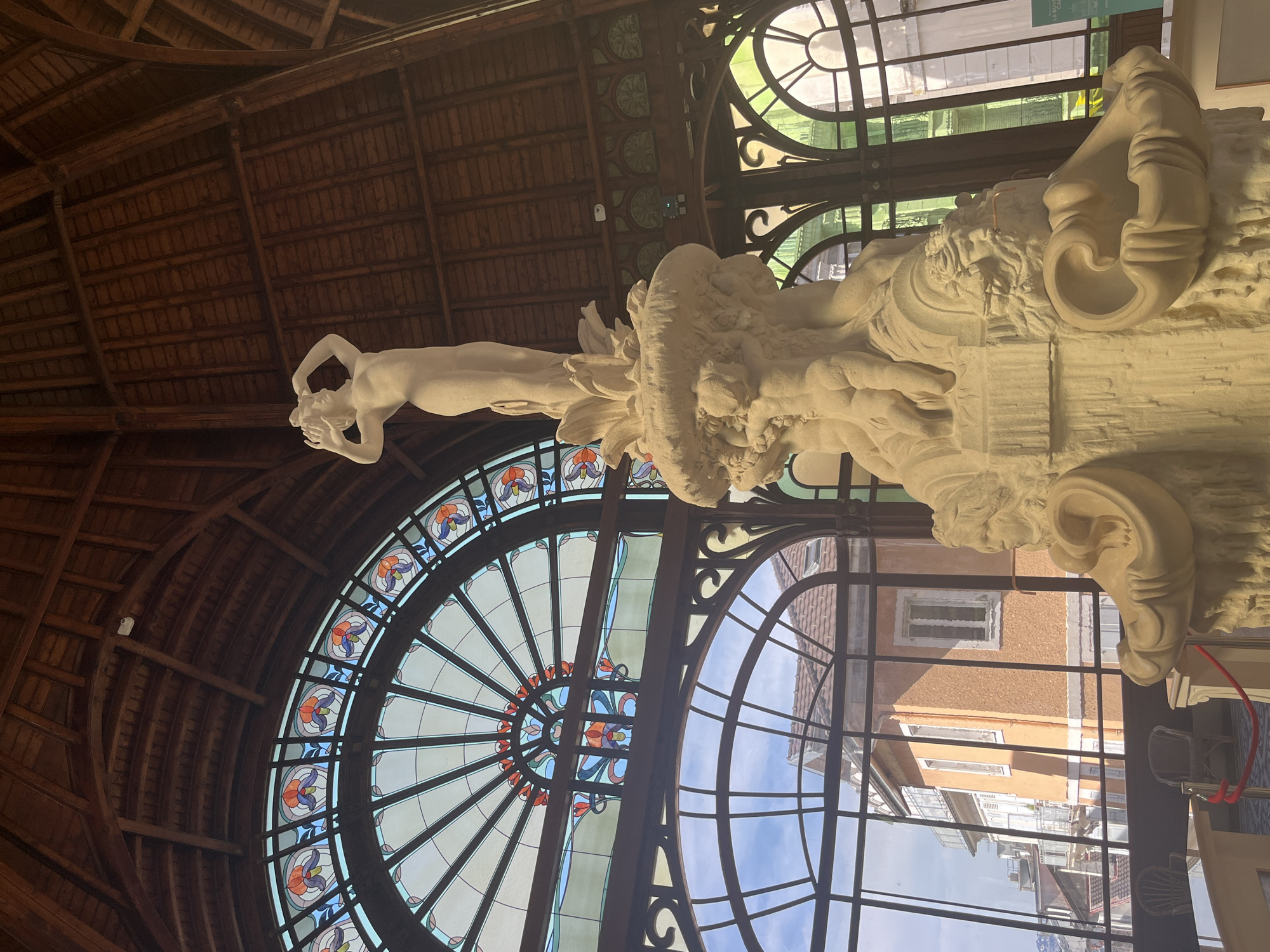The image depicts the interior of the Palais Lumière in Évian-les-Bains, France. The focal point is a large, ornate fountain sculpture made of light-colored stone. The fountain features a nude female figure at the top, with cherubic figures and architectural elements below. The sculpture is set within a grand hall with a vaulted wooden ceiling and large, arched windows. One of the windows is a stained-glass panel with floral motifs in shades of blue, orange, and purple. Another large window offers a view of the exterior, showing buildings with orange and white facades, rooftops, and a bright sky. In the foreground, a red rope barrier separates the viewer from the fountain. Two wicker chairs and a table are visible through the window. The overall scene suggests an indoor public space, likely a museum or historical building, characterized by ornate architecture and a central artistic installation.