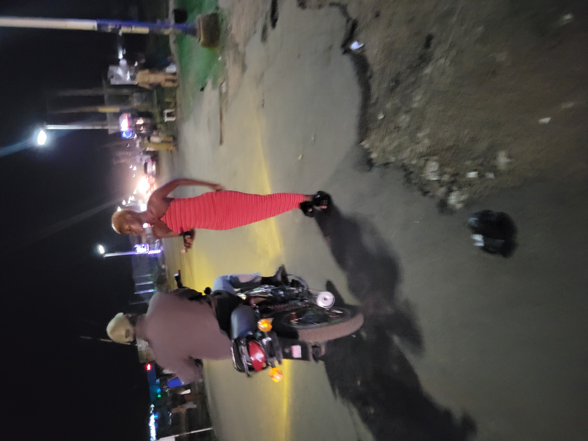 A nighttime outdoor shot shows a person in a form-fitting red dress standing near a motorcycle on a paved surface. The person has light-colored hair and is looking to the left. Next to them, a person is sitting on a motorcycle, facing to the right. To the right of the motorcycle, there is a dark helmet lying on the ground. In the background, there are lights and what appear to be buildings or structures, typical of a city street at night. The location is Unknown, Unknown.