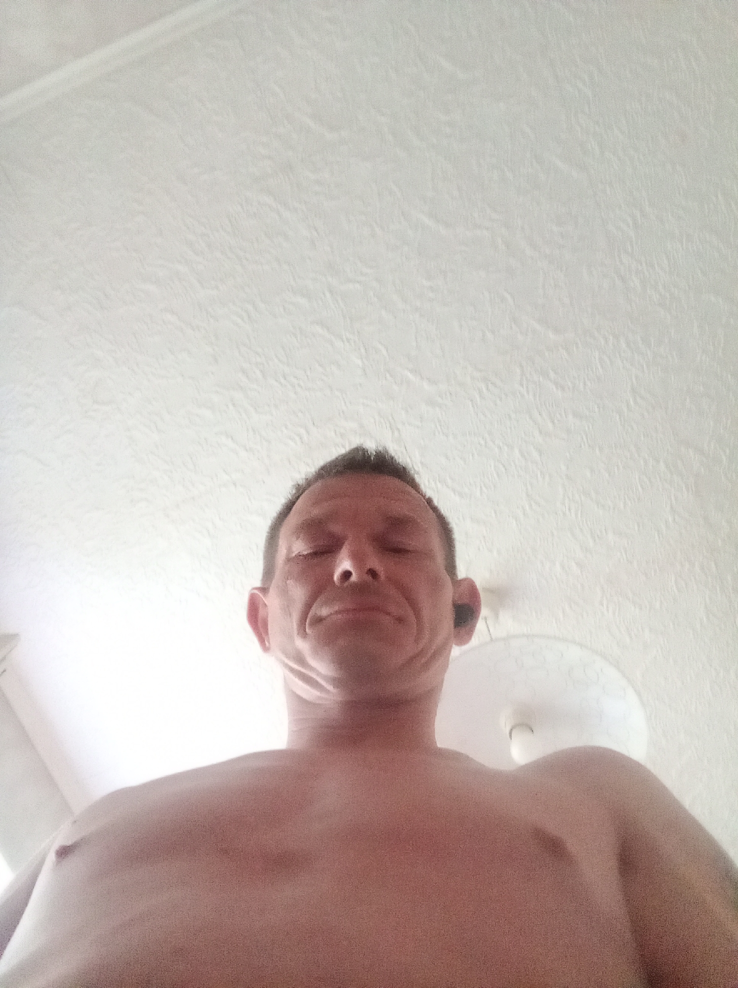 The image shows a low-angle, close-up shot of a bare-chested individual, likely male, from the shoulders up. The person's eyes are closed, and they appear to be smiling slightly. They have short, dark hair and are wearing a black earbud in their right ear. The background is a textured white ceiling, with a light fixture visible towards the right side of the frame. The lighting suggests an indoor setting, possibly a home. There are no discernible landmarks or contextual clues that would place this image specifically in Tapa, Estonia.