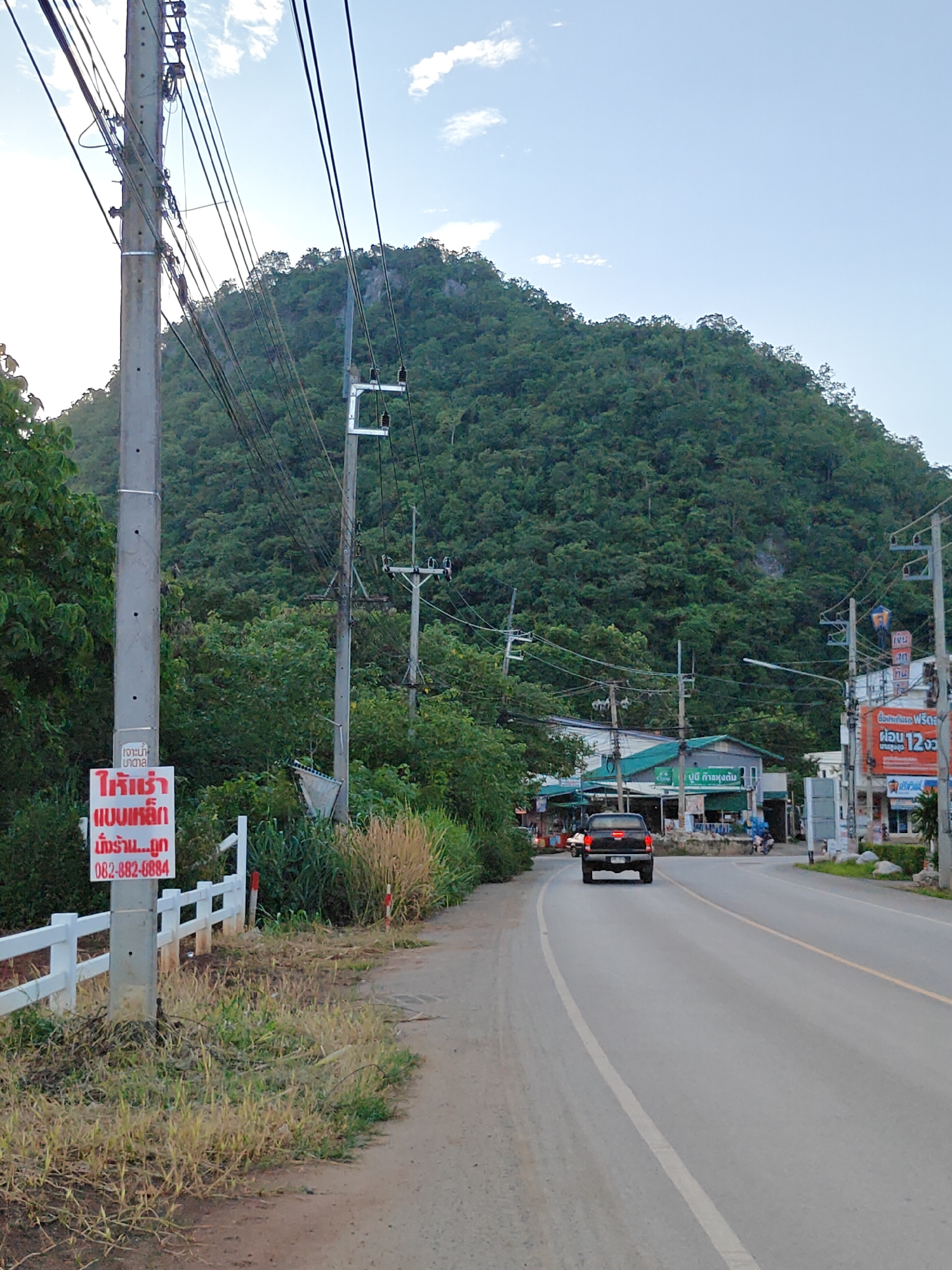 The image shows a road in Mu Si, Thailand, with a dense, tree-covered mountain in the background. A utility pole with numerous wires stands on the left side of the road, next to a white fence. A red and white sign is attached to the utility pole, featuring Thai script. A black pickup truck is driving away from the camera on the paved road, which has yellow and white lines marking the lanes. Several buildings, some with awnings and signage, are visible on the right side of the road, indicating a commercial area. A motorcycle is parked near one of the buildings. The sky is partly cloudy, and the overall scene suggests an outdoor environment in a mountainous region.