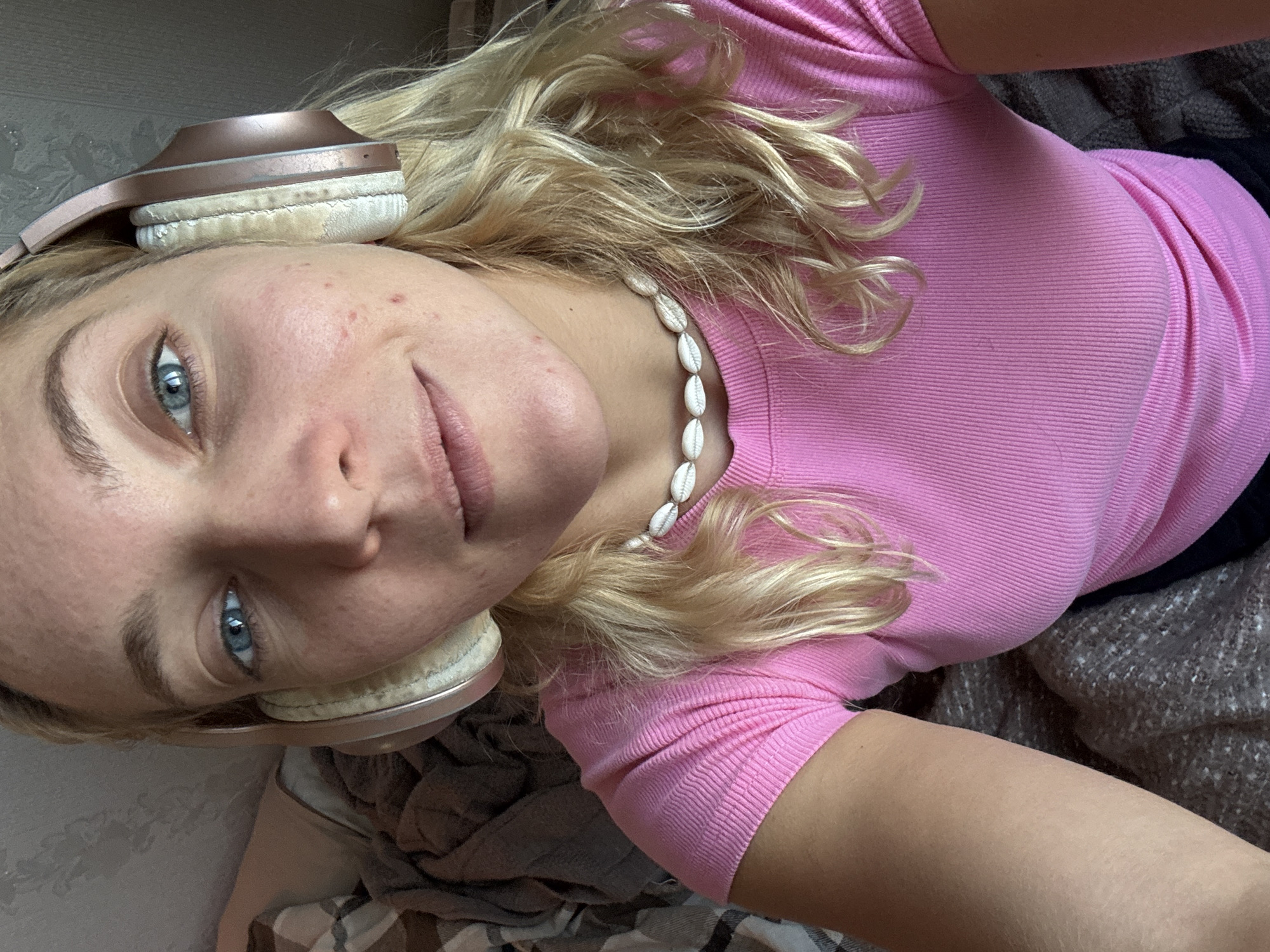 A young woman with wavy, blonde hair and striking blue eyes is depicted in a close-up, top-down selfie. Her head is tilted to the left, and she looks directly at the camera with a calm, slightly smiling expression. She is wearing a ribbed, short-sleeved pink top.
Notable accessories include a pair of rose gold or copper-colored over-ear headphones, which are resting on her head. The white earcups of the headphones show signs of wear and slight discoloration. Around her neck, she wears a white necklace made of cowrie shells.
Her skin has some small red marks or acne visible on her forehead and cheeks. The background suggests an indoor setting, possibly a bed or couch, with a patterned, dark grey and brown blanket or fabric visible. A light-colored wall with a subtle pattern can be seen behind her head. The lighting is soft and ambient, indicative of an indoor environment. There is no visible text in the image. This scene takes place in Minsk, Belarus.