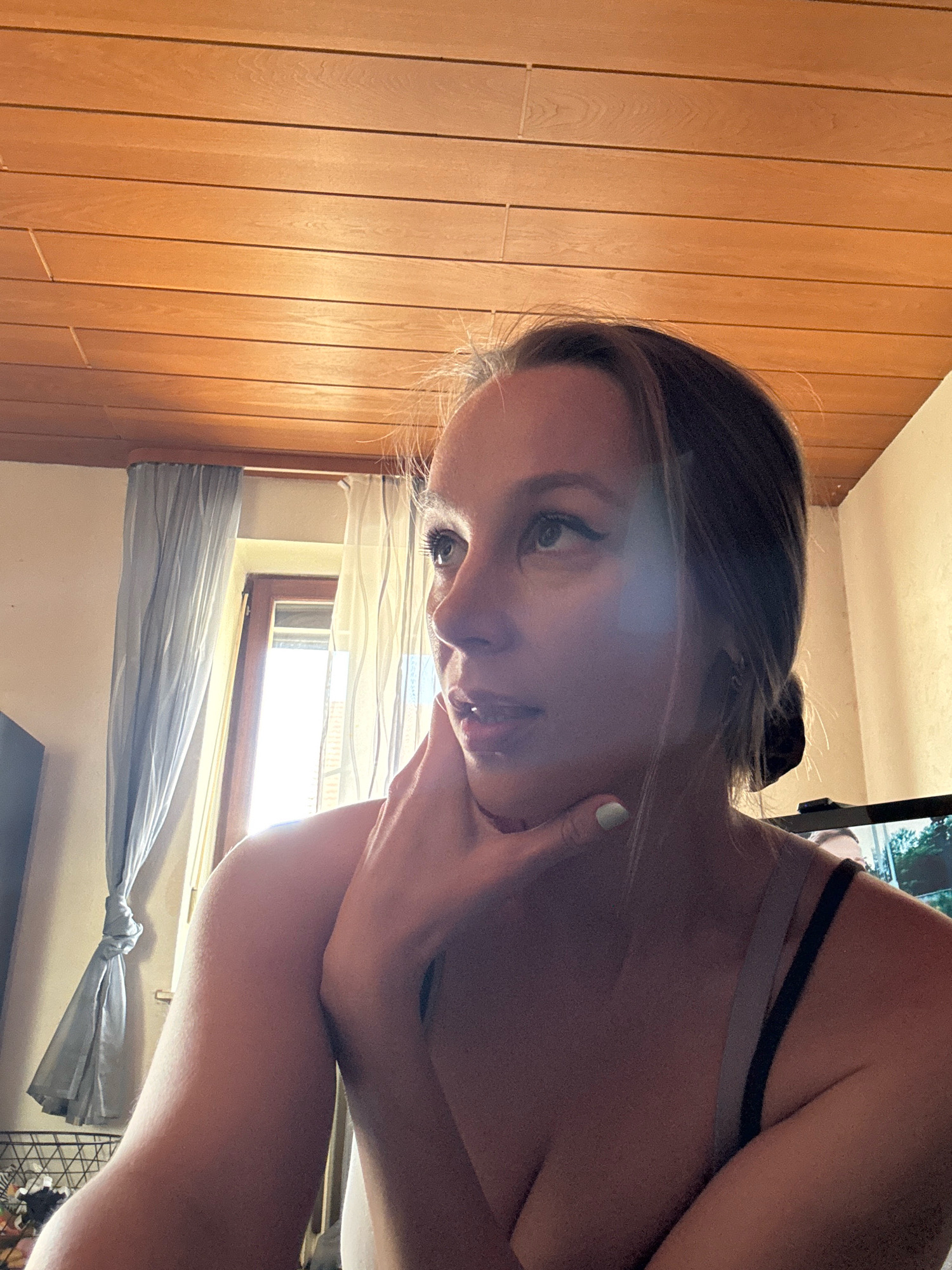 The image shows a close-up, medium-angle shot of a young woman from the chest up, positioned indoors. She is looking upwards and to her right, with her left hand resting on her chin. Her hair is pulled back, and she is wearing a bra. The background features a wooden paneled ceiling, a window with light-colored curtains, and a computer monitor. The scene appears to be within a residential setting in Überherrn, Germany, based on the provided location context. The lighting suggests it is daytime, with natural light coming from the window.
