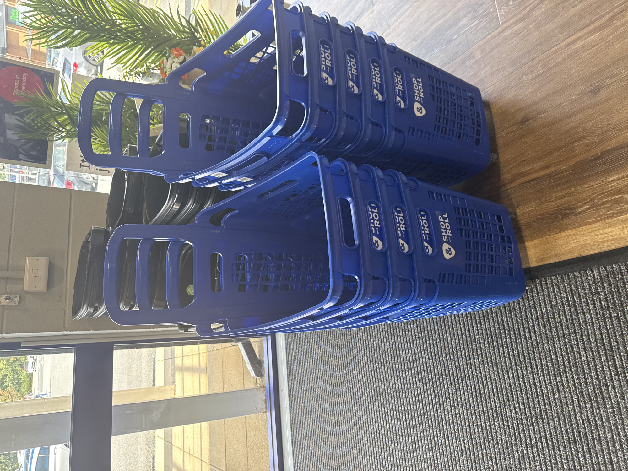 The image shows a stack of blue plastic shopping baskets with the "SHOP and ROLL" logo. The baskets are located indoors, near a doorway that leads to an outdoor area with a gray ribbed mat on the floor. In the background, through the doorway, a parking lot with cars and some trees are visible, suggesting the location might be a retail store or a similar establishment. There is also some artificial foliage and a sign that reads "Donate in store today" visible in the background. The scene is well-lit, with the primary focus on the shopping baskets. There are no people clearly visible in the image. The shopping baskets are neatly stacked and appear to be ready for use. The overall environment suggests a retail or service setting. The location in Darlington, United Kingdom is not explicitly evident from the image itself but is provided as context.