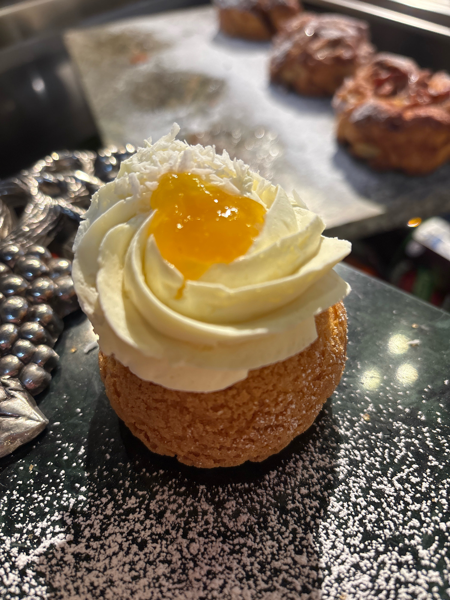 A single, round pastry topped with white frosting and a dollop of orange jam sits on a dark, speckled surface dusted with powdered sugar. The pastry has a rough, golden-brown exterior. To the left of the pastry, a decorative, silver object with a grape-like pattern is partially visible. In the blurred background, several more pastries can be seen on a light-colored surface. The lighting suggests an indoor setting, possibly a bakery or cafe. The scene does not contain any recognizable landmarks or elements that would indicate a specific location in Riga, Latvia.