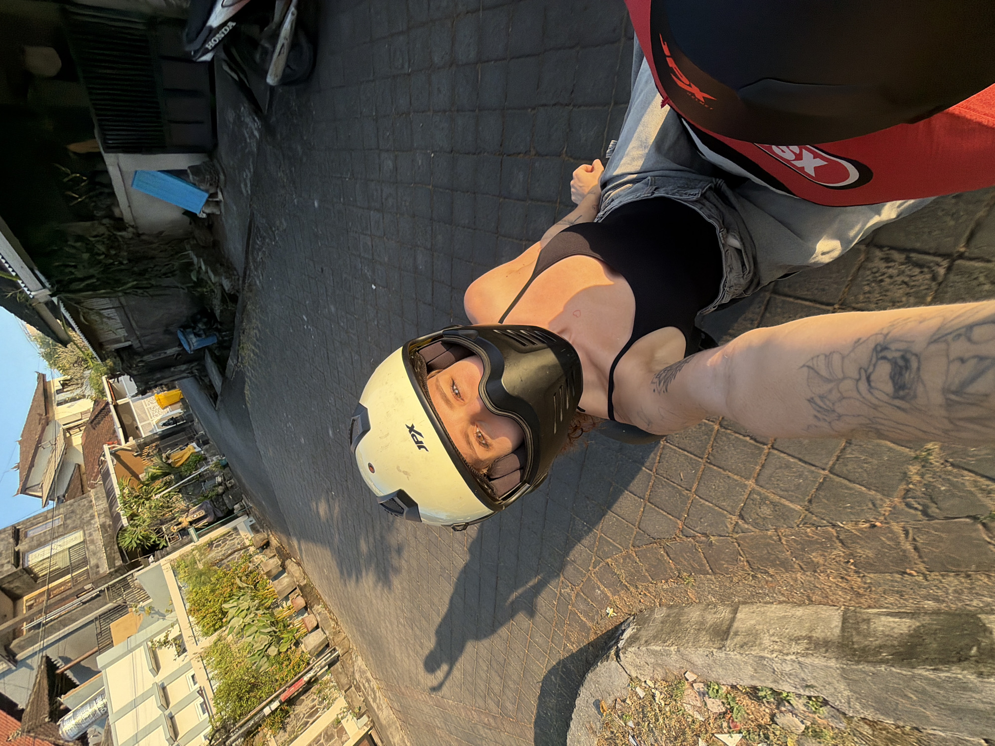 A point-of-view shot captures a woman wearing a pale yellow helmet and black face mask, her eyes visible through the visor. She is leaning back on a motorcycle, with her arm extended holding the camera. She wears a black tank top and grey jeans. A shadow of her raising her arm is cast on the stone-paved road. Buildings with red tile roofs and stone walls line the street, with lush green plants growing between them. A parked motorcycle with "HONDA" written on it is visible in the background. The scene is set in a narrow street in North Kuta, Indonesia, bathed in the warm glow of the setting sun.
