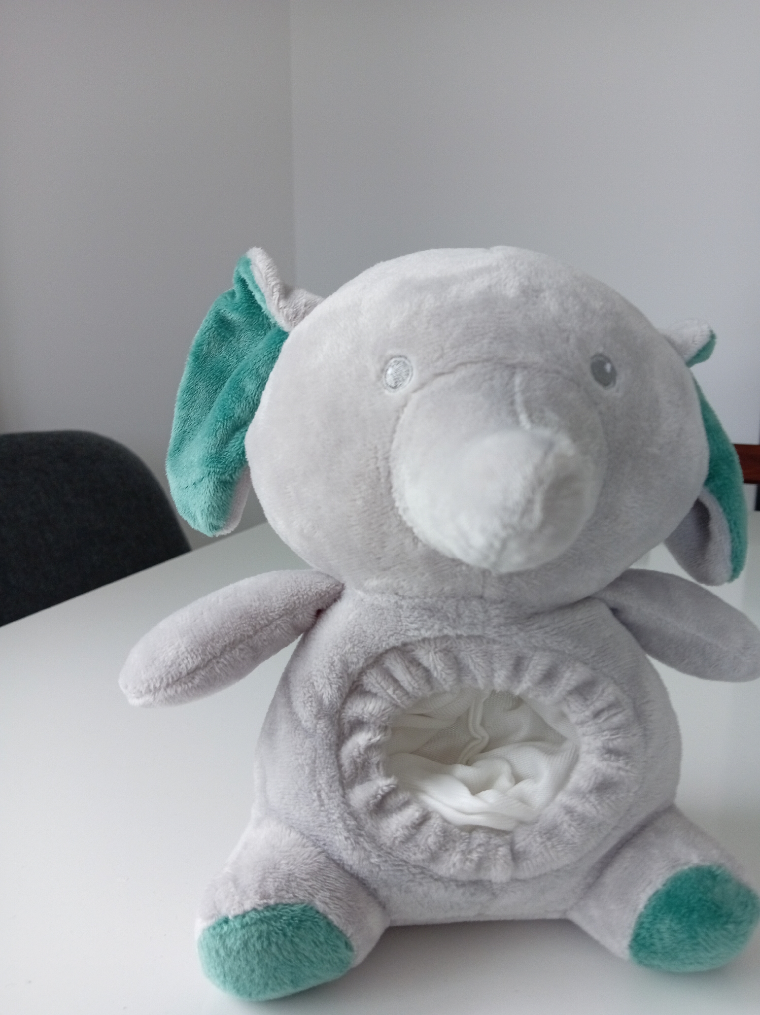 A close-up shot features a plush elephant toy sitting on a white surface, likely a table. The elephant is gray with teal accents on its ears and feet. It has two round, gray eyes, and its trunk is extended forward, open at the tip, revealing a white lining inside. The belly of the elephant also has an opening, stuffed with white fabric. In the background, a dark gray chair is partially visible, and a plain, light gray wall occupies the upper portion of the frame. The lighting is soft and diffused, with no discernible light sources. The scene appears to be indoors, in a sparsely decorated environment.