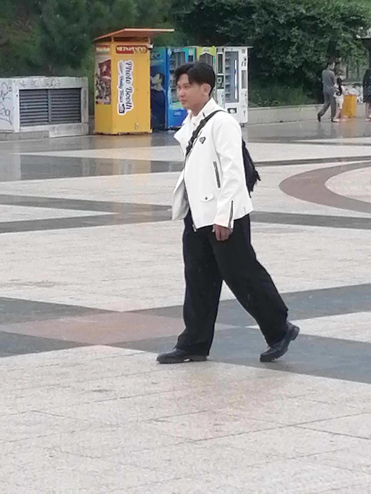 A man is walking through a public square. He is wearing a white jacket and black pants. He has a black backpack on his shoulder. There are several vending machines and a photo booth in the background. The ground is paved with stone tiles. The image appears to be taken on a cloudy day. The man is walking away from the camera and towards the right side of the frame. The overall scene is unremarkable. The location of Da Lat, Vietnam is not evident from the image.