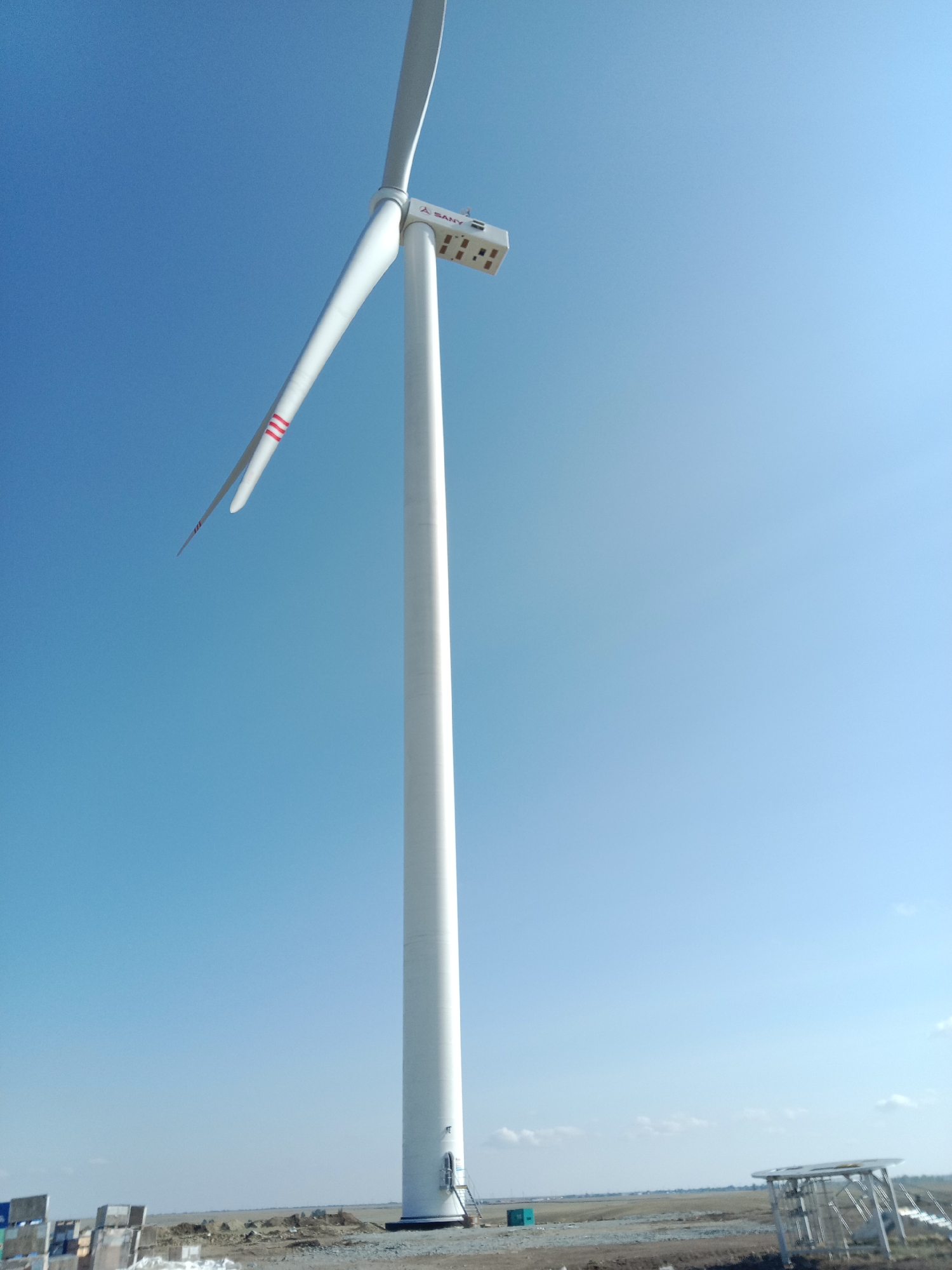 This is a low-angle, full-shot photograph of a large white wind turbine against a clear blue sky. The wind turbine is positioned in the foreground, dominating the left side of the frame. Its tall, cylindrical tower is white, with a closed door visible near the base. A ladder extends from the ground to the door. The turbine's blades are angled upwards and to the left. The larger blade has three red horizontal stripes near its tip. The nacelle, or housing at the top of the tower, is white and rectangular with some visible equipment and a logo that appears to read "SANY".
In the background, the landscape is flat and barren, with some signs of construction or industrial activity. To the left of the turbine, there are several stacked crates or building materials, some with blue and white markings. Further into the background, a small teal-colored structure is visible. To the right and further back, a metal frame structure with a translucent roof is present, possibly part of a solar panel installation or another piece of industrial equipment. The horizon line is low, with a faint line of low-lying clouds or distant structures visible. The overall scene suggests an open, possibly industrial or construction site under a bright, clear sky. The location is specified as Akzhar, Kazakhstan.