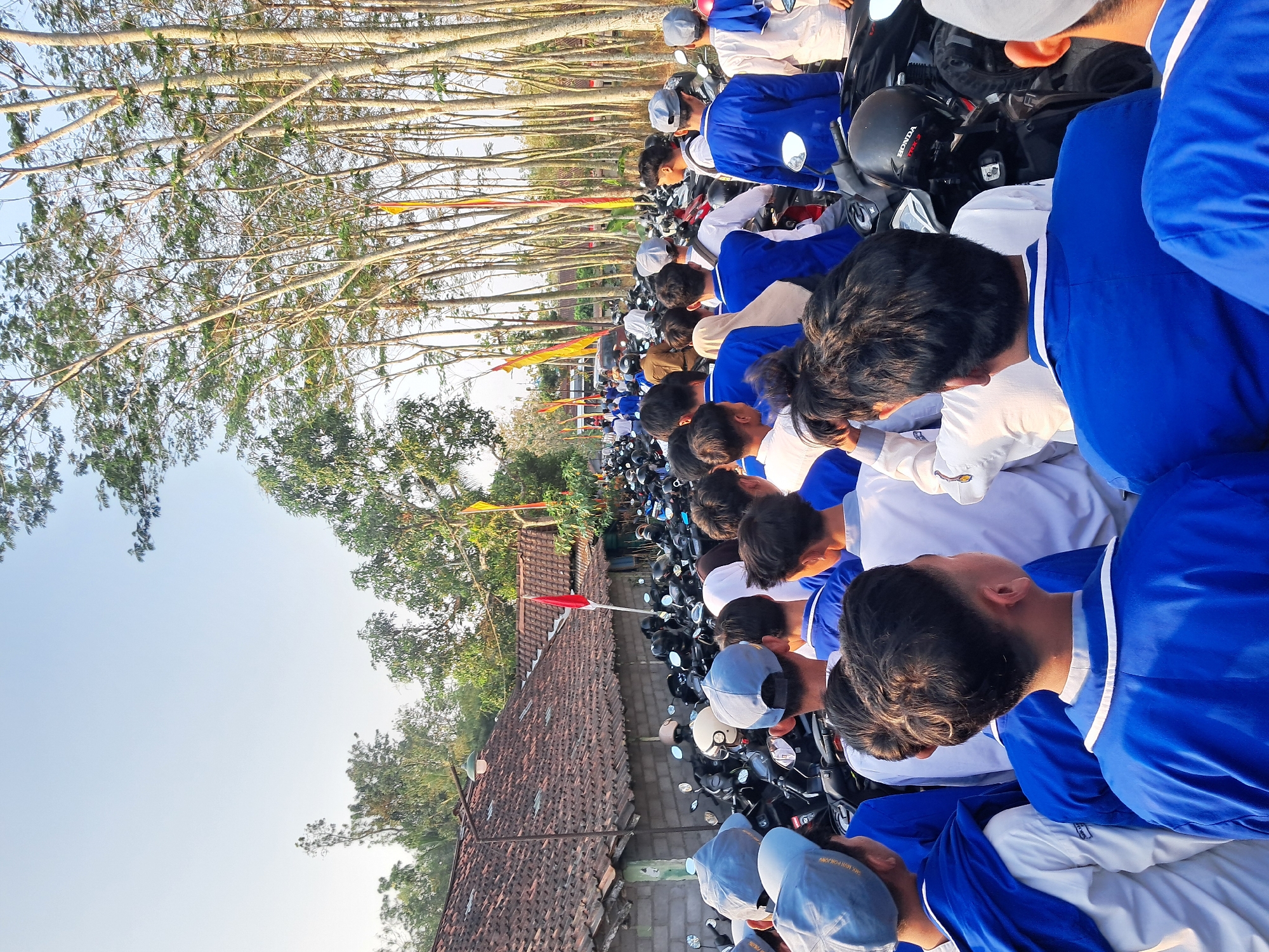 The image depicts a street scene in Ponjong, Indonesia, characterized by a large gathering of people, primarily young individuals, many of whom are wearing blue uniforms and caps. They are standing amidst a densely parked area of motorcycles, suggesting a communal event or transportation hub. The background features traditional Indonesian architecture with terracotta roofs, a brick wall, and lush vegetation including tall trees lining the street. Red and yellow flags are visible, adding a festive or ceremonial element to the setting. The sky is clear and bright, indicating it is daytime, likely late afternoon given the angle of the sunlight. The overall impression is of an organized assembly, possibly related to a school or community activity, taking place in a rural or semi-urban Indonesian environment.