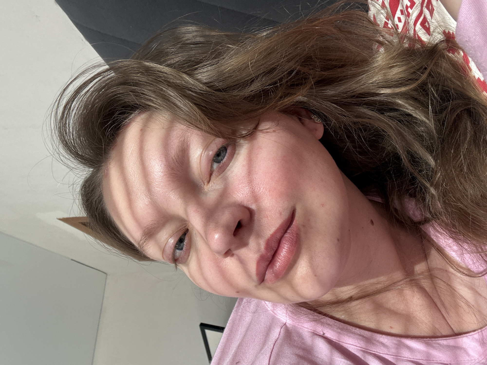 A close-up, angled shot of a person's face with light and shadow lines across it. The person has light skin, blue eyes, and light brown hair. They are wearing a light pink shirt. The background includes parts of a dark, possibly black or gray object, and a patterned cloth. There is no information provided in the image to indicate the location is Izhevsk, Russia.