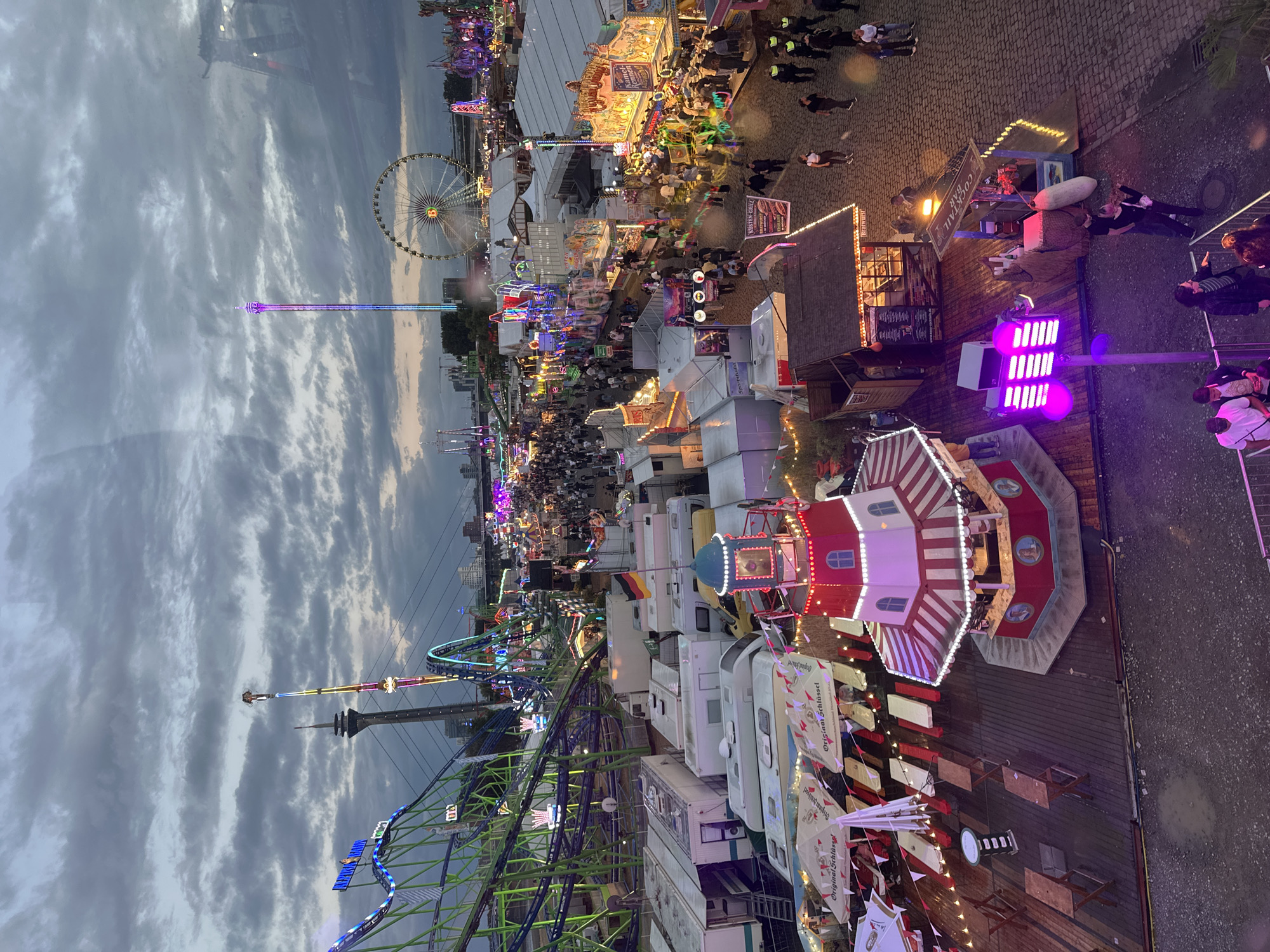 The image captures a panoramic view of a bustling funfair set up along the Rhine embankment in Dusseldorf, Germany. The sky is overcast with a mixture of gray and pale blue clouds, suggesting it is either early evening or late afternoon. The funfair is illuminated by numerous colorful lights from various rides and attractions, including a prominent Ferris wheel in the mid-ground and a tall, illuminated tower ride. A green roller coaster is visible on the left side of the frame, with its tracks winding into the distance. Numerous white caravans or mobile homes are parked in a row, possibly housing vendors or staff. In the foreground, there's a red and white striped kiosk with a lighthouse-like structure on top, decorated with lights. Wooden tables and benches are set up around this kiosk and under white and red umbrellas, indicating an outdoor seating area for food or drinks.
A dense crowd of people are seen walking through the fairgrounds, browsing stalls, and enjoying the attractions. Some of the stalls display signage for food and drinks, such as "KÜSTEN-GRILL" and "Brathurst," and one is labeled "COCKTAIL BAR." German flags are visible on some of the structures. The ground is paved with cobblestones in some areas and wooden decking in others. The overall atmosphere suggests a lively and vibrant event, with the city's skyline and a bridge faintly visible in the background, grounding the scene in Dusseldorf.