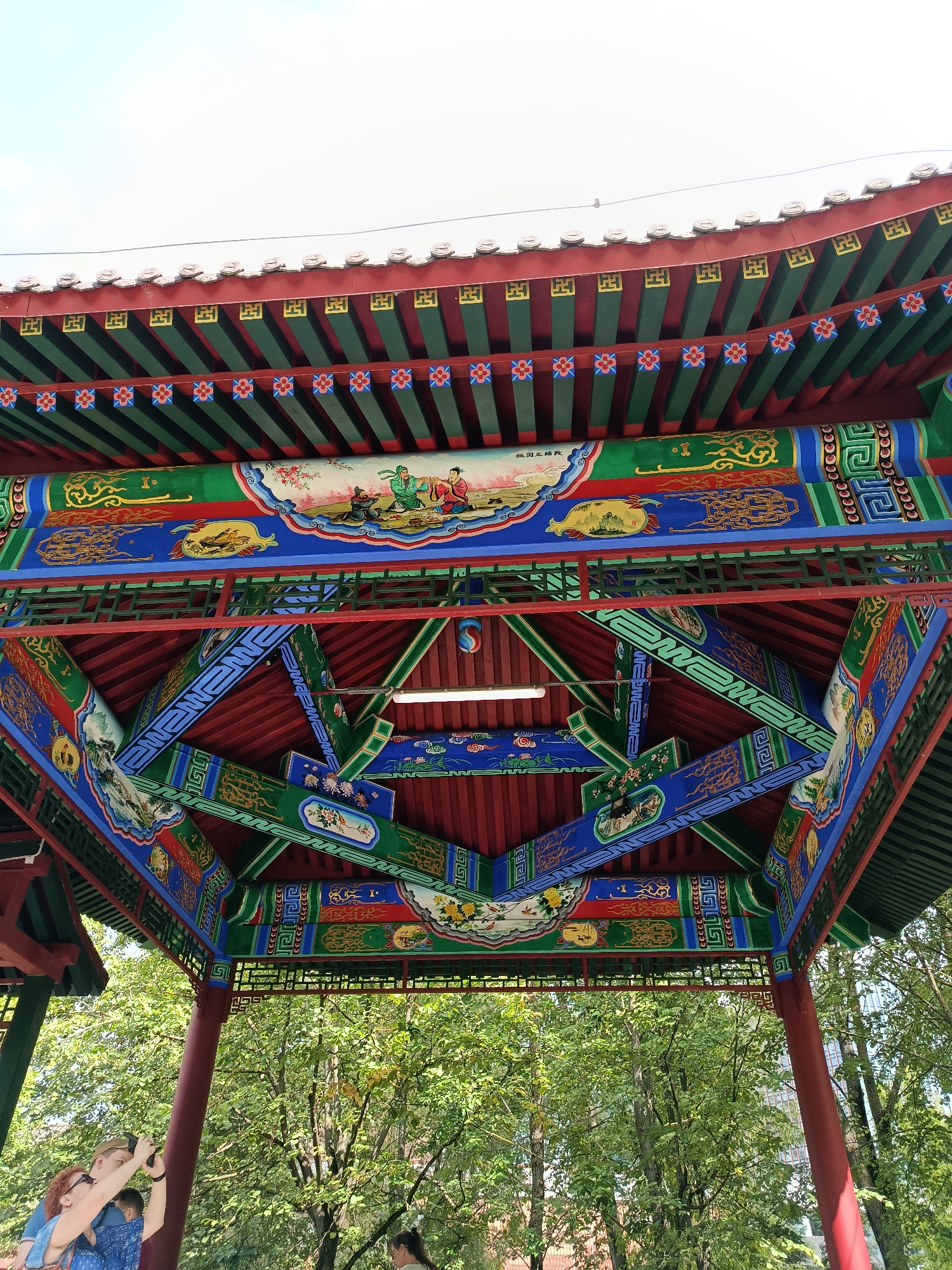 The image shows a close-up, upward view of the ornate ceiling of a traditional Chinese-style pavilion, possibly located in Moscow, Russia, given the context provided. The pavilion's roof and beams are extensively decorated with vibrant colors, including red, green, blue, and yellow. Intricate patterns and motifs are painted throughout, featuring stylized clouds, floral designs, and depictions of figures in traditional attire, suggesting scenes from Chinese art or mythology. The underlying structure consists of exposed red beams and green rafters, creating a complex geometric framework.
In the lower part of the image, the tops of green trees are visible, indicating an outdoor setting with lush foliage. A few individuals are present in the foreground. On the left, a person with reddish-colored hair, wearing a blue top, is holding up a smartphone to take a photograph of the pavilion ceiling. Next to them, another person, presumably a man, is also holding a phone. Further to the right, a person with dark hair tied back is partially visible, looking upwards. The sky above is a pale, overcast white.