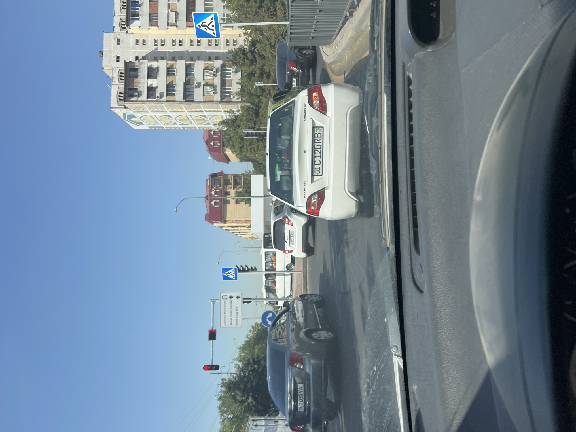 This image captures a view from inside a vehicle in a traffic situation on a sunny day in Tashkent, Uzbekistan. In the foreground, the dashboard and steering wheel of the vehicle are visible. The middle ground displays a street scene with several cars, including a gray Chevrolet sedan in the left lane and a white Chevrolet sedan in the right lane. A bus is seen in the distance, and other vehicles are also present in the traffic. Traffic lights, street signs, and pedestrian crossing signs are visible. Apartment buildings and trees line the street, indicating an urban environment. The sky is clear and blue. The license plate of the gray Chevrolet is visible and reads "01 T 030 YA". The license plate of the white Chevrolet is also visible and reads "01 C 120 RB".