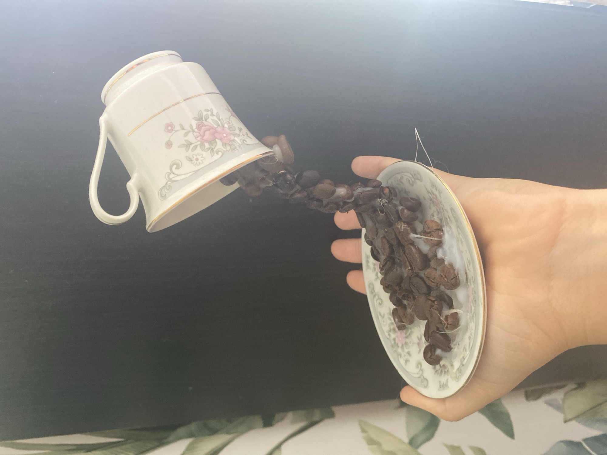 A hand is holding a small, ornate saucer containing coffee beans. A teacup, also ornate, is tipped over above the saucer, appearing to pour the coffee beans into it. The teacup is white with floral designs and gold trim. The coffee beans are dark brown and some appear to be connected by thin white threads, creating the illusion of a pour. The background is a dark, slightly textured surface, possibly a wall, with a hint of greenery from what appears to be a tapestry or wallpaper on the left side. The lighting is bright, coming from the right side of the image, creating a glare. The scene is set indoors, with no identifiable city or geographical markers present in the image.