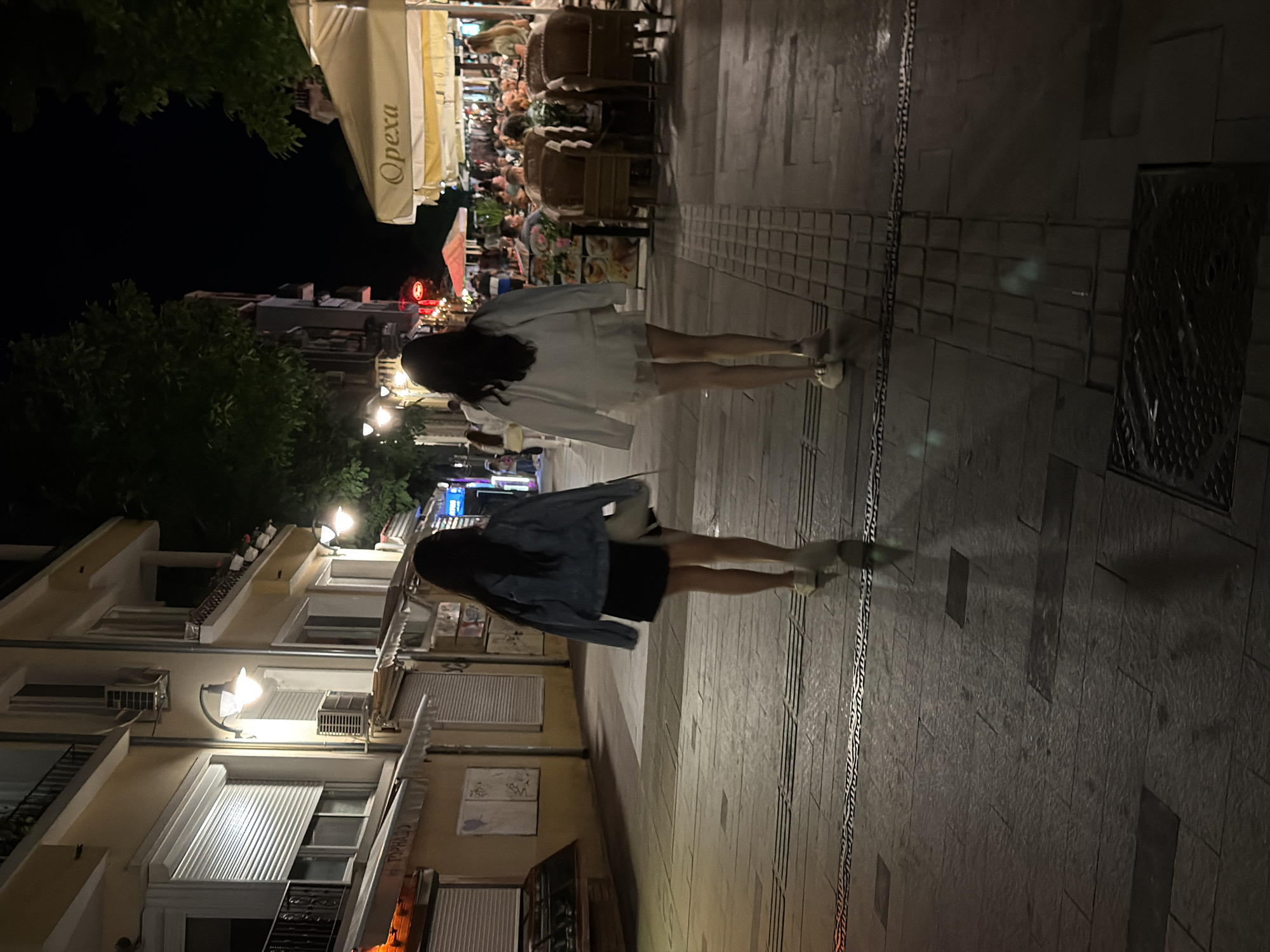 The media file shows a street scene at night in Burgas, Bulgaria. Two women are walking away from the camera, one in front of the other. The woman in the foreground is wearing a denim jacket over a black dress and white sneakers. The woman behind her is wearing a white jacket over a white dress and white sneakers. Both women have dark hair and appear to be walking on a paved sidewalk.
In the background, there are buildings with awnings and lights, suggesting a commercial area with restaurants or shops. There are also trees and outdoor seating visible, indicating a lively atmosphere. The lighting suggests it is evening or nighttime, with streetlights and interior lights illuminating the scene. The cobblestone pavement and architectural style of the buildings provide clues to the location. One awning has text that appears to read "Opexa". The overall mood is casual and relaxed, with the women enjoying an evening stroll.