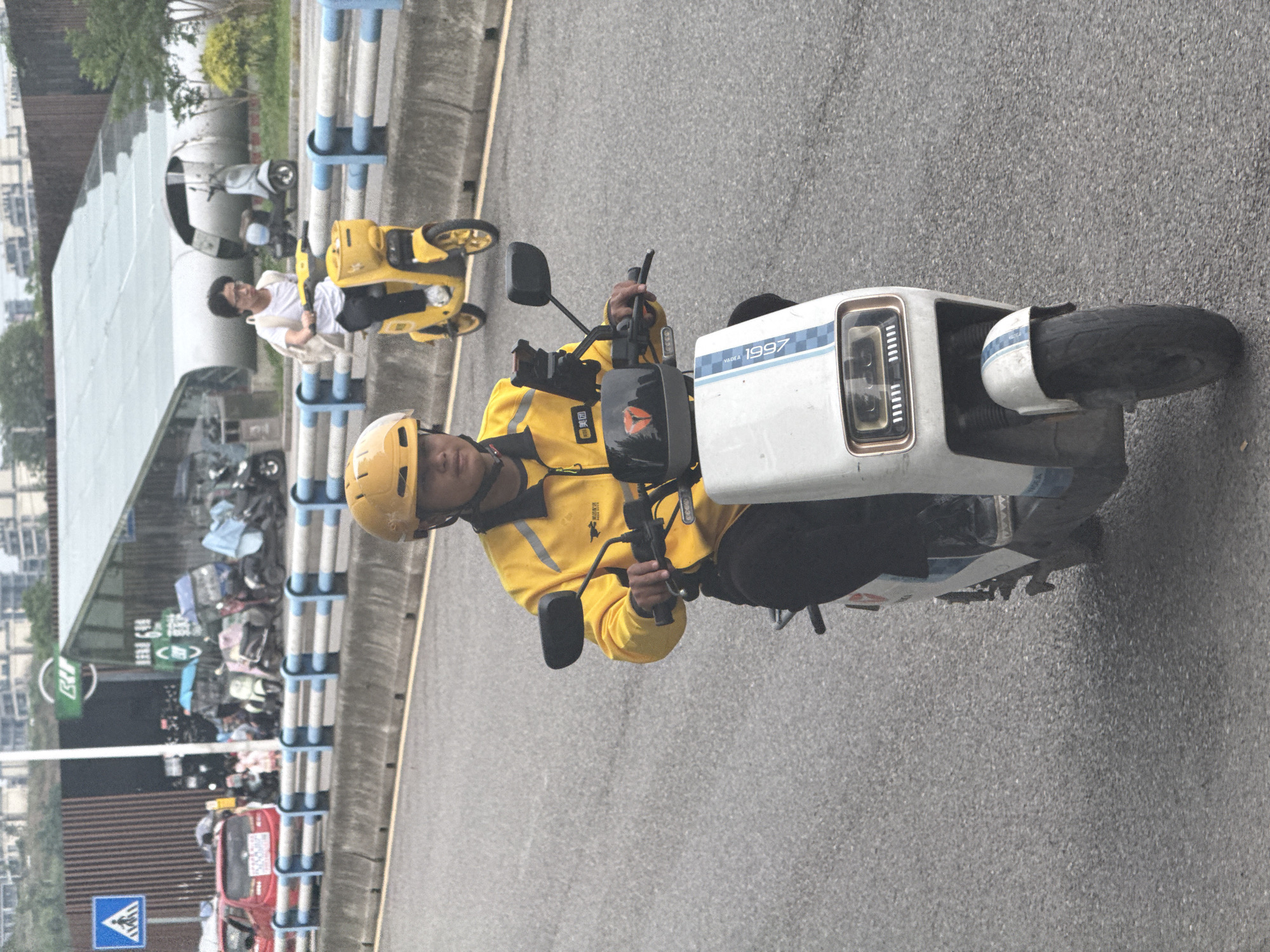 A medium-shot, ground-level, eye-level photograph shows a man on an electric motorcycle in the Liangjiang, China area. The man, wearing a yellow helmet and a yellow jacket with a black logo, is the primary subject. He appears to be operating the motorcycle, with his hands on the controls. The motorcycle is predominantly white with blue accents and the text "YADEA 1997" on the front.
In the background, there is a sheltered parking area with several yellow scooters parked within it, including one being occupied by a person in a white shirt and black pants. A blue and white striped barrier runs along the edge of the road. Further in the background, there is a red vehicle, a green sign, and various other parked motorcycles. Buildings and greenery can be seen in the distance. The ground is paved with asphalt.