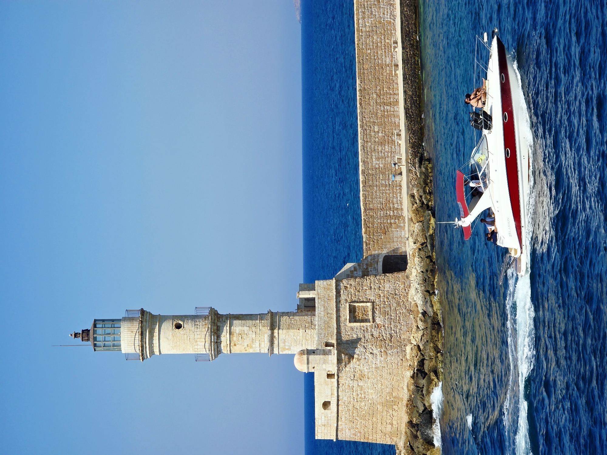A historic lighthouse stands tall on a stone pier, with a clear blue sky overhead. The lighthouse is a cylindrical structure made of weathered stone, with a lantern room at the top. A white and red motorboat speeds across the deep blue sea in the foreground, leaving a white wake. Two people are visible on the boat, one lying down and the other sitting. In the background, a long stone wall extends from the base of the lighthouse. This scene is the Venetian Lighthouse of Chania, a prominent landmark located at the entrance of the old Venetian harbor in Chania, Crete, Greece.