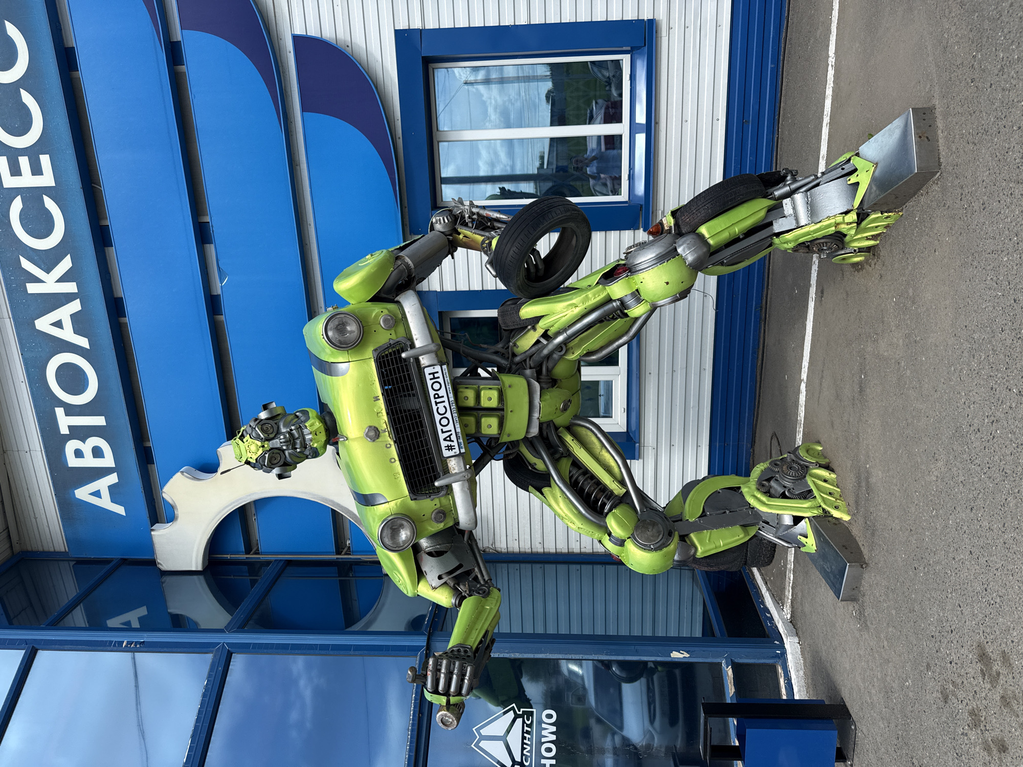 An outdoor shot depicts a large, bright green, metal robot sculpture standing in front of a building. The robot is constructed from various car parts, including headlights, wheels, and springs. It has a license plate on its chest that reads "# АГОСТРОН". In the background, the building has large blue signage with white text that partially reads "АВТОАКСЕС". The building also features windows with blue frames. To the left of the robot, a glass-fronted building entrance with a logo that includes the word "HOWO" is visible. The ground is asphalt, and a blue trash bin is located to the left of the robot. The scene appears to be a commercial area, possibly a car service or parts store, in the city of Izhevsk, Russia. The lighting suggests it is daytime.
