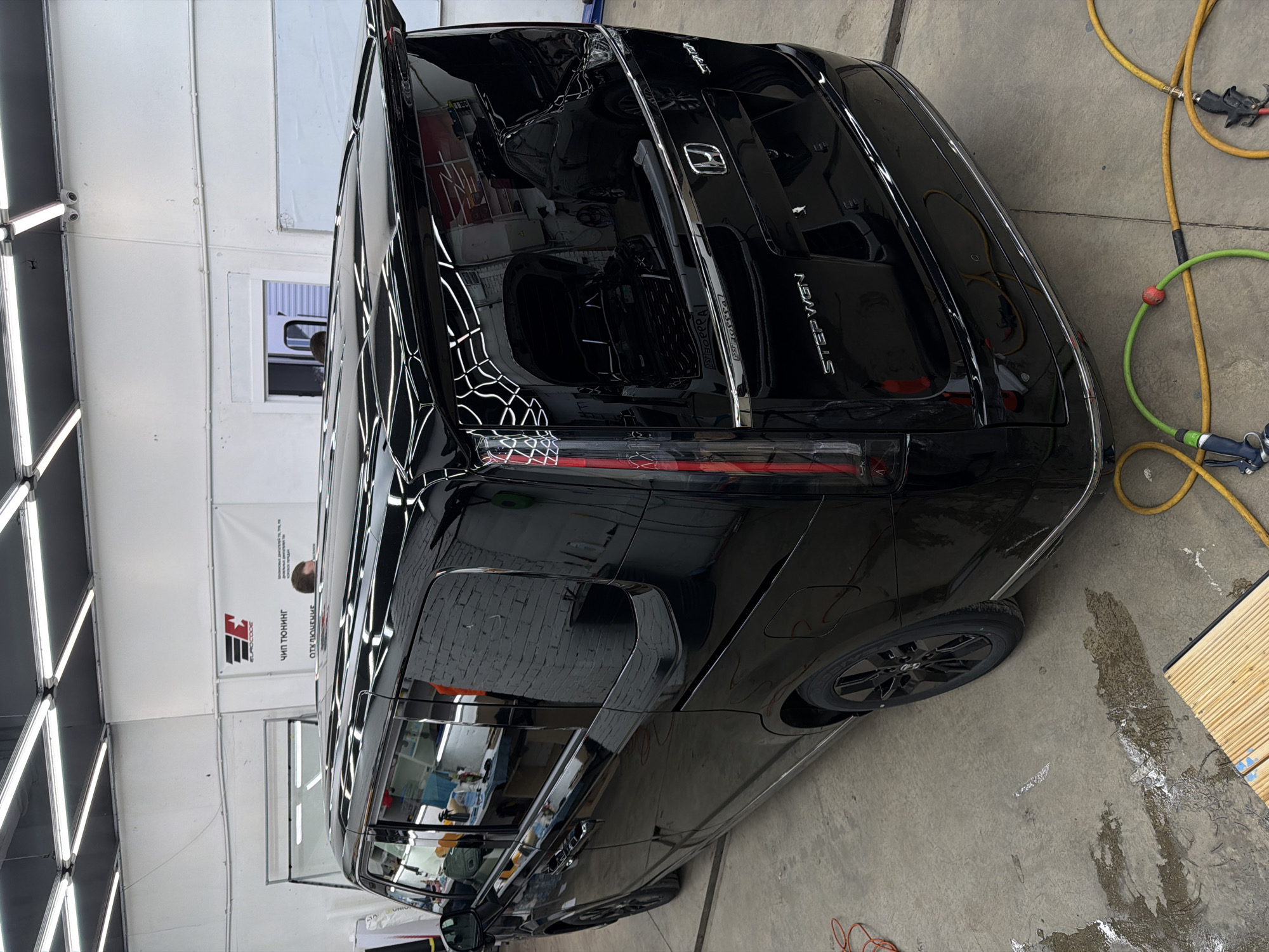 A black Honda Stepwgn is in a car detailing shop in Krasnoyarsk, Russia, evidenced by the "EuRoCode" signage. The car is shiny and appears to have been recently detailed. Various hoses and tools are visible on the concrete floor. A person is visible through the side window of the car. The shop is well-lit with fluorescent lights on the wall and ceiling.
