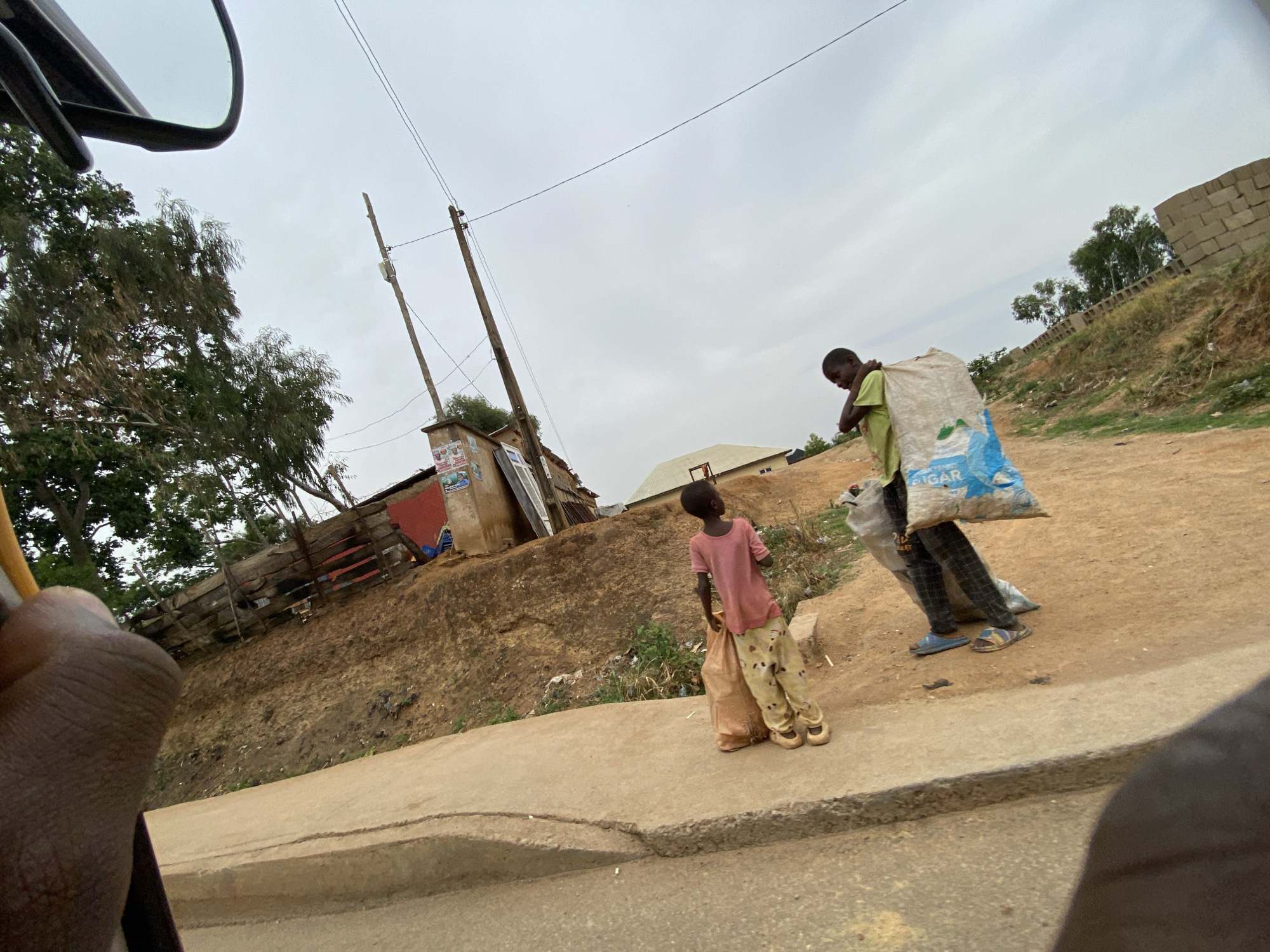 This photograph depicts a street scene in Bukuru, Nigeria, under an overcast sky. The image is taken from a low angle, possibly from inside a vehicle, with a dark object (likely a mirror or vehicle frame) partially visible in the upper left and a hand in the lower left.
In the foreground, a concrete curb separates a dirt path or road from the viewer's position. On this path, two individuals are prominent. An adult or older boy, wearing a light green t-shirt, dark trousers, and sandals, carries a large white and blue sack on his back, supported by his shoulder. The sack clearly bears the word "SUGAR" printed on it. Walking beside him, a young child, dressed in a pink t-shirt and light-colored patterned trousers with sandals, holds a smaller, light brown bag.
The background shows an upward sloping terrain, a mix of bare earth and some green vegetation. On this higher ground, several rudimentary structures are visible, including a small building with a red wall and another with what appears to be corrugated metal siding. Wooden utility poles with power lines crisscross the sky. Further back, more trees and some uncompleted block walls or structures can be seen. The sky is uniformly gray and cloudy, indicating an overcast day. The scene portrays daily life with individuals transporting goods in what appears to be a semi-urban or rural setting.