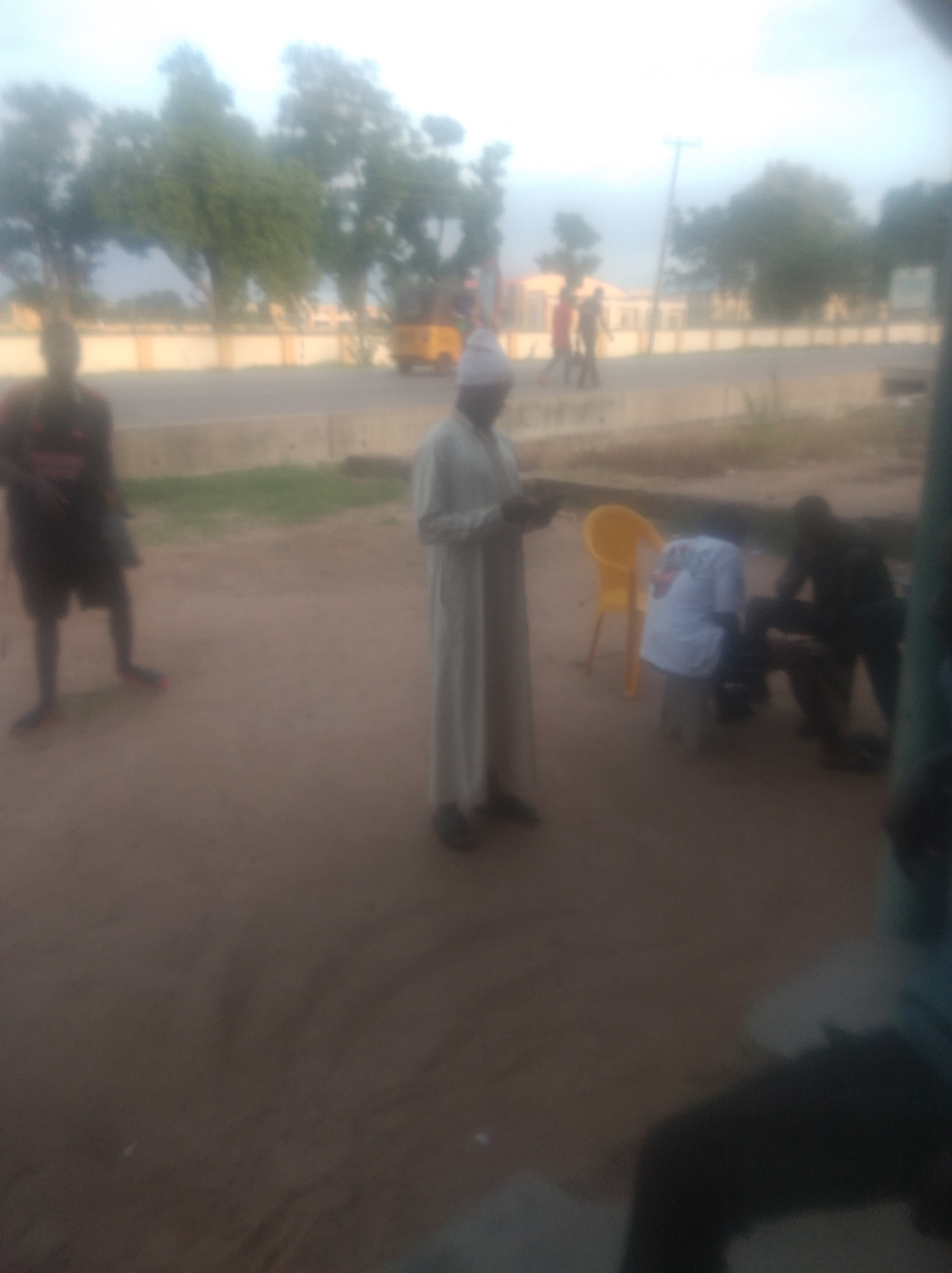 The image depicts an outdoor scene, likely in Jalingo, Nigeria, as indicated. In the foreground, several individuals are present. A person in a light-colored robe stands centrally, holding a dark object in their hands. To the left, another person is visible from the waist down, wearing dark shorts and a dark shirt with red accents. To the right, two individuals are situated near a yellow plastic chair. One is wearing a white shirt, and the other, dressed in dark clothing, is kneeling or bending over. In the background, a road is visible, bordered by a beige wall and trees. A yellow three-wheeled vehicle, possibly a tuk-tuk, is also present on the road. The sky is overcast.
