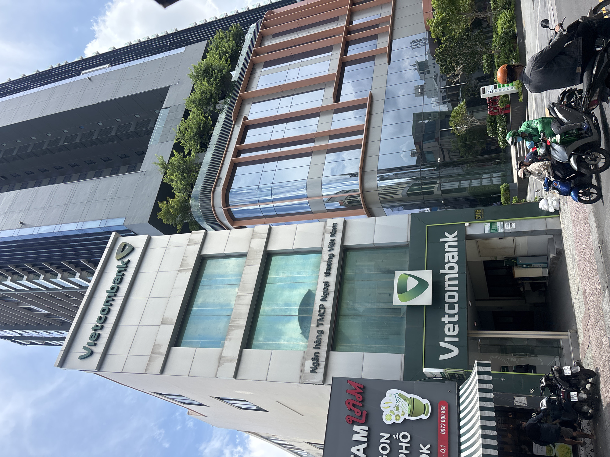 In an urban outdoor scene under a cloudy sky, a modern bank building with the "Vietcombank" logo is prominently featured. The building is white and has large glass windows. Next to it, a taller, darker building with a more complex facade, featuring vertical brown accents and multiple floors of glass windows, rises into the sky. Green trees are visible on a terrace of the taller building. In the foreground, a street scene unfolds with several motorcycles and people. Two individuals on a motorcycle wear green and blue masks and helmets. Another person on a motorcycle wears an orange helmet. A sign for "TAM LAM" with an image of a mortar and pestle with lime and chili is visible on the left. The ground is paved with textured tiles, and a sidewalk runs alongside the street.