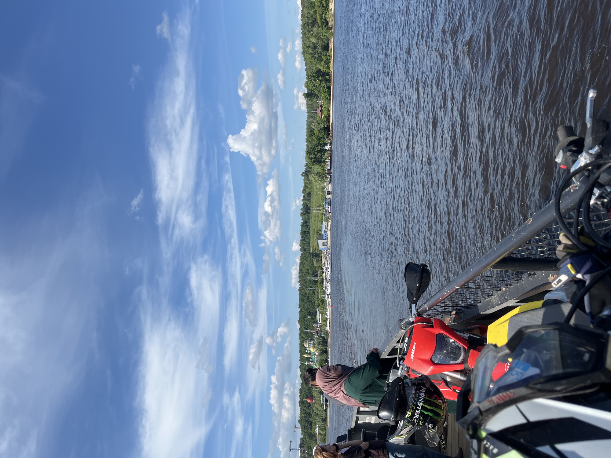 The image depicts a bright, sunny day aboard a ferry traversing a wide body of water, likely the Kazanka River in Kazan, Russia, given the background structures. The sky is a clear blue with scattered white, wispy clouds.
In the distance, the shoreline of Kazan is visible, characterized by a dense, vibrant green tree line. Above the trees on the mid-left, a construction crane and partially visible buildings indicate urban development. Toward the center-right background, distinct colorful buildings with onion-shaped domes, characteristic of the Kazan Riviera Aquapark or a nearby cultural complex, rise above the trees. Further along the shore to the right, a marina with numerous white boats and yachts is docked, accompanied by a sandy beach area and other small structures.
In the foreground, on the deck of the ferry, several off-road or enduro motorcycles are parked, facing generally towards the right. A prominent red motorcycle, bearing "DHL" branding on its front fairing, is visible in the mid-foreground. A black helmet adorned with a "Monster Energy" logo rests on or near the handlebars of this red bike. Partially visible in the extreme foreground is the handlebars and blurred helmet of another motorcycle, yellow and black in color. A dark metal railing separates the motorcycles and passengers from the dark, rippled water, which exhibits small waves from the movement of the ferry or wind.
On the left side of the frame, two individuals are leaning against the railing, observing the distant shore. One woman, positioned centrally on the left, wears a dark green long-sleeved top and a light pink headscarf (hijab), looking towards the right with a gentle smile. Another person is partially obscured behind her. A third individual, with visible red hair and wearing a dark top, is partially seen on the far left. The scene captures a moment of transit, with passengers and their motorcycles enjoying the view of the city's waterfront.