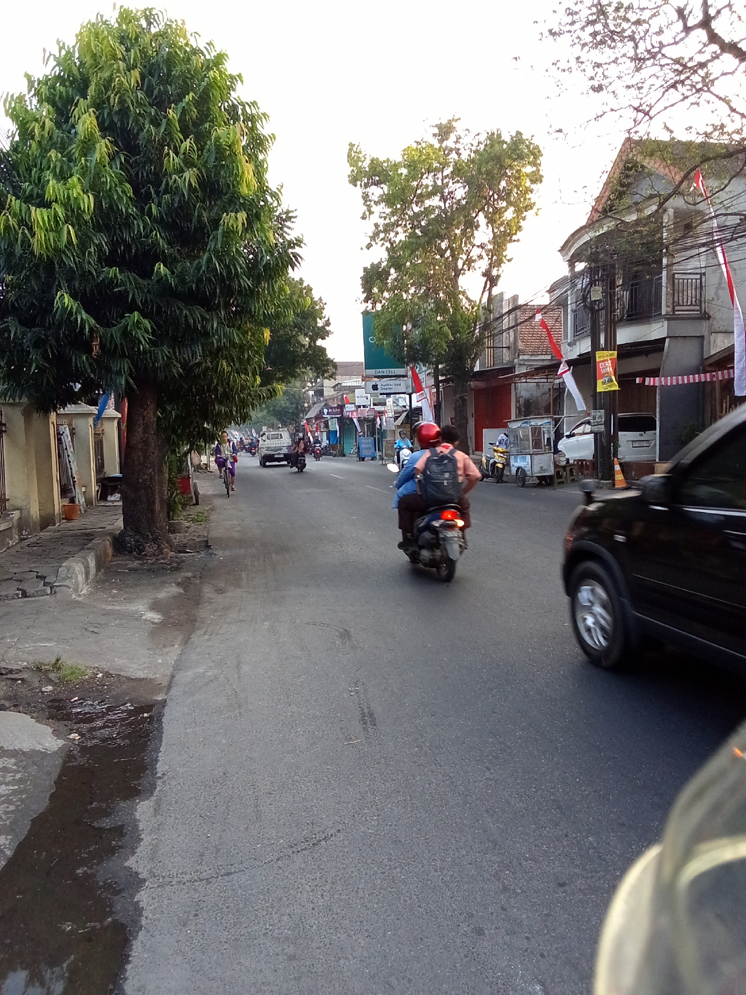 A street scene in Kediri City, Indonesia, captures a bright day with a focus on a motorcycle carrying two passengers. The motorcycle is black with a red license plate and is moving down a paved road. The passenger in the rear is wearing a red helmet and a dark backpack, while the driver wears a light-colored shirt. In the foreground, the right side of a black SUV is visible. The left side of the road features a sidewalk and a large, leafy tree. Buildings line the right side of the street, some adorned with Indonesian flags, suggesting a festive or patriotic occasion. Other vehicles, including bicycles and another motorcycle, are present further down the road. The overall impression is a bustling urban environment.