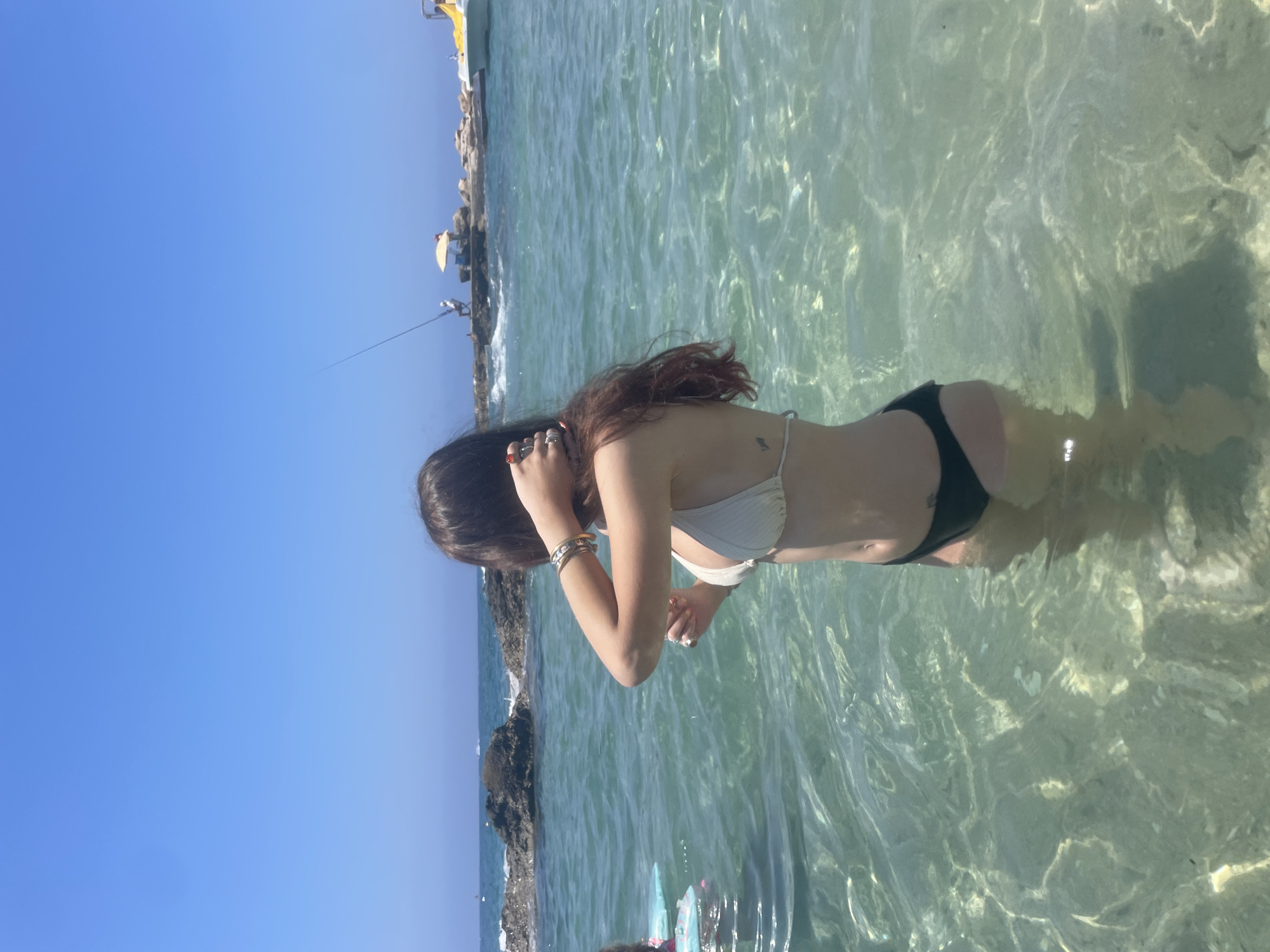 The image depicts a sunny day at a coastal location, likely in Paphos, Cyprus. In the foreground, a young woman stands in clear, shallow water, facing away from the camera. She has long, dark hair and is wearing a white bikini top and dark bottoms. She is touching her hair with one hand and appears to be adjusting it. Her left arm is adorned with bracelets. In the background, the ocean stretches to the horizon under a clear blue sky. Rocks are visible near the shore, and a person is fishing from a rocky outcrop. Further in the distance, a yellow boat is docked. The water is transparent, revealing the sandy seabed beneath. The scene conveys a sense of leisure and enjoyment of a summer day by the sea.