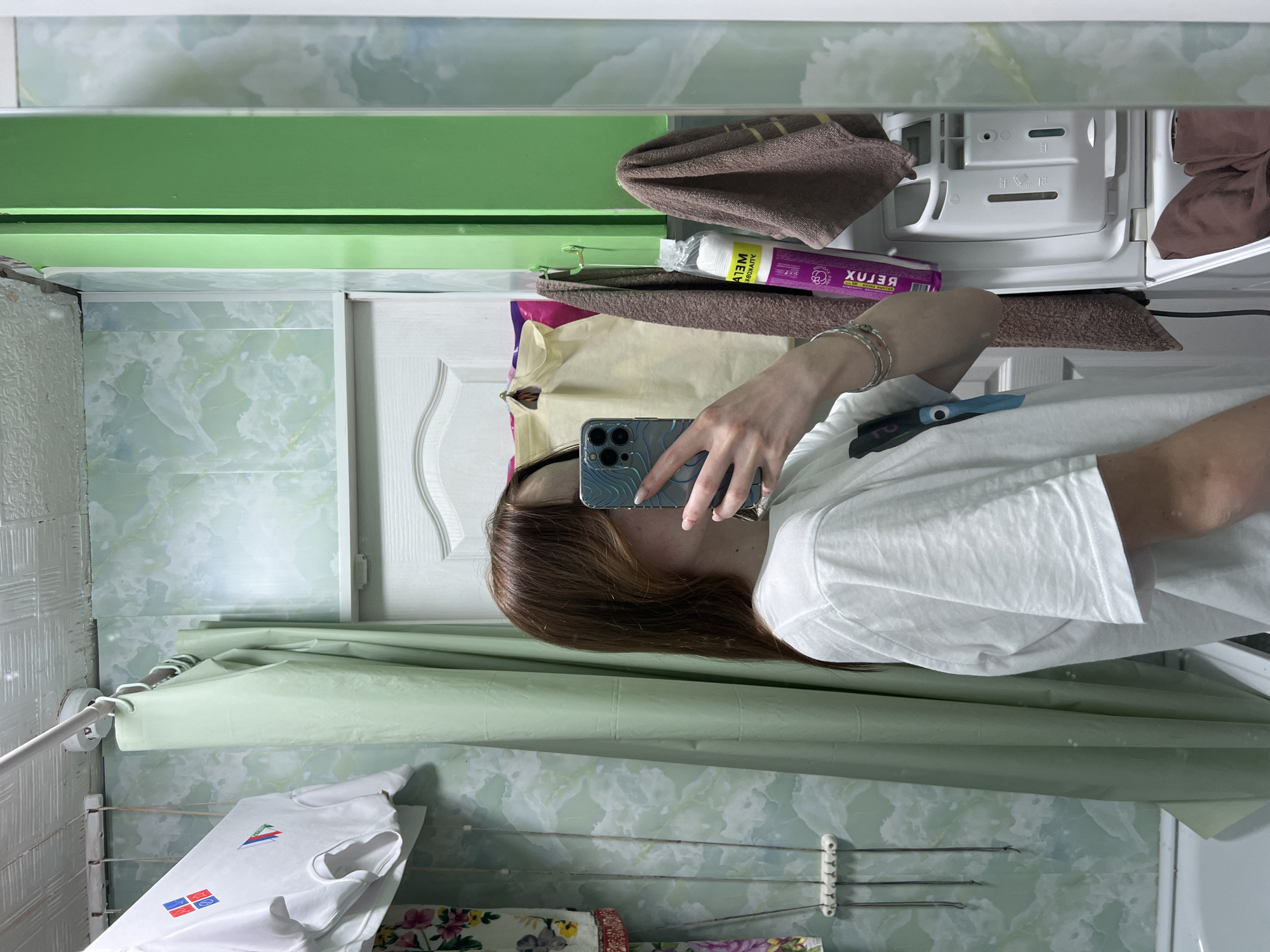 A person is taking a selfie in a bathroom in what appears to be an apartment. The person has long brown hair and is wearing a white t-shirt. They are holding a smartphone. A green shower curtain hangs in the background. There is a clothes drying rack with clothing. The bathroom has green and white patterned tiles. Above the person and clothing rack is a white cabinet with brown towels and purple shampoo or lotion. The city is Chaikovsky, Russia.