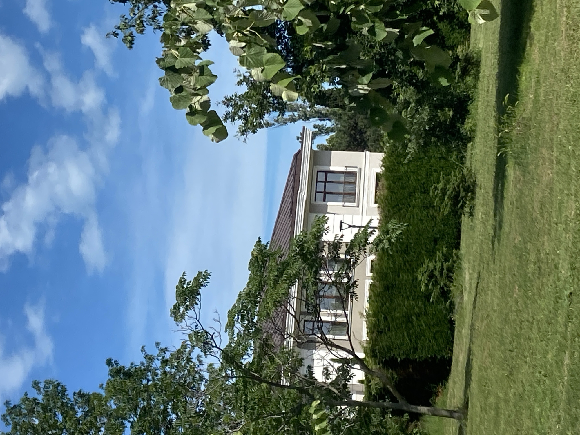 The image shows a modern, two-story house with a brown roof, white walls, and large windows. It is surrounded by lush green landscaping, including a manicured hedge in front of the house and mature trees framing the scene. A lamppost stands near the hedge. The sky is a clear blue with scattered white clouds. The image appears to be taken on a sunny day. There are no people or animals visible in the image. The architectural style suggests a suburban or semi-rural setting. Given the context provided, this house is located in the city of Varna, Bulgaria.