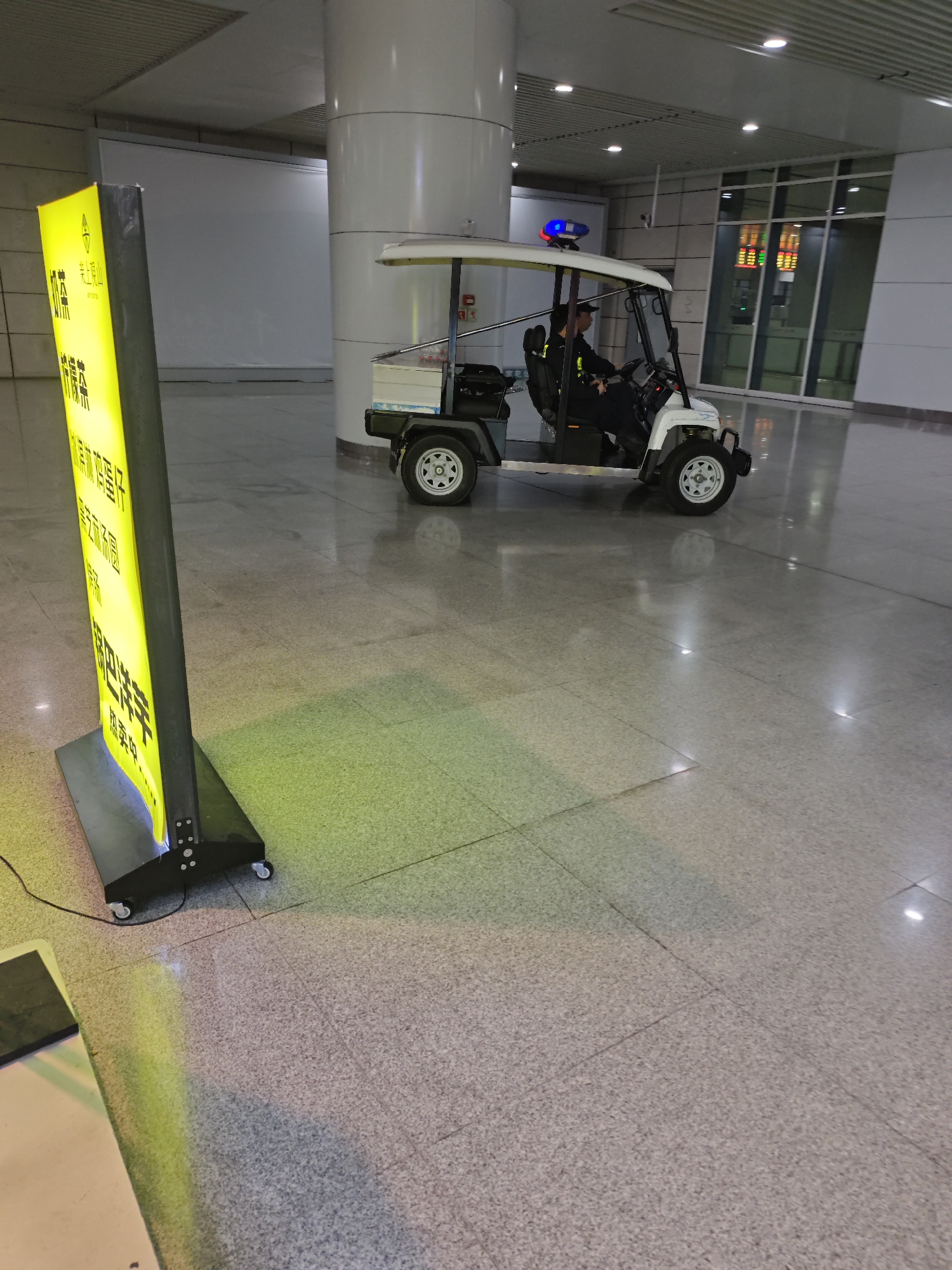 The image shows the interior of a building, possibly an airport terminal or convention center. A white electric cart with a driver in uniform is situated near a large pillar. A yellow sign is placed in the foreground, with what appears to be Chinese text on it. The floor is tiled. Given the information, the location is somewhere in the Liangjiang New Area of Chongqing, China.