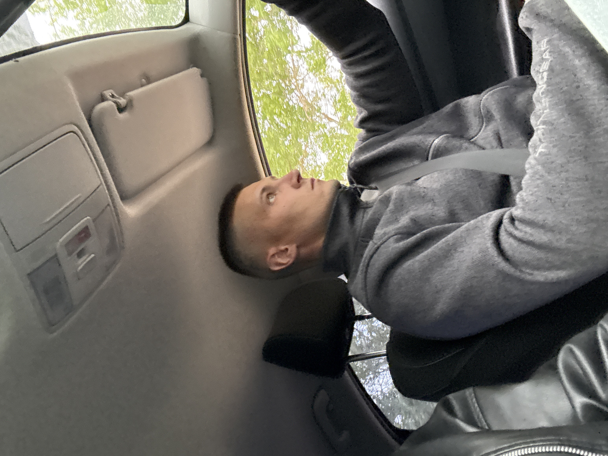 A male individual, appearing to be in his 20s or 30s, with a short fade haircut, is seated in the front passenger position of a vehicle in Chaikovsky, Russia. He is wearing a dark grey, textured zip-up fleece jacket and has a light grey seatbelt secured across his chest. His right arm is extended forward. He is looking to his right, out the side window, at bright green tree foliage, indicating an outdoor, possibly daylight, setting. The car's interior features a light grey headliner, an overhead console with integrated lighting, and a passenger-side sun visor. A black leather seat with visible stitching is partially visible in the lower left foreground, and a black headrest is discernible behind the subject. White text, partially legible as "YPMNGEAR", is embroidered on the sleeve of his left arm.