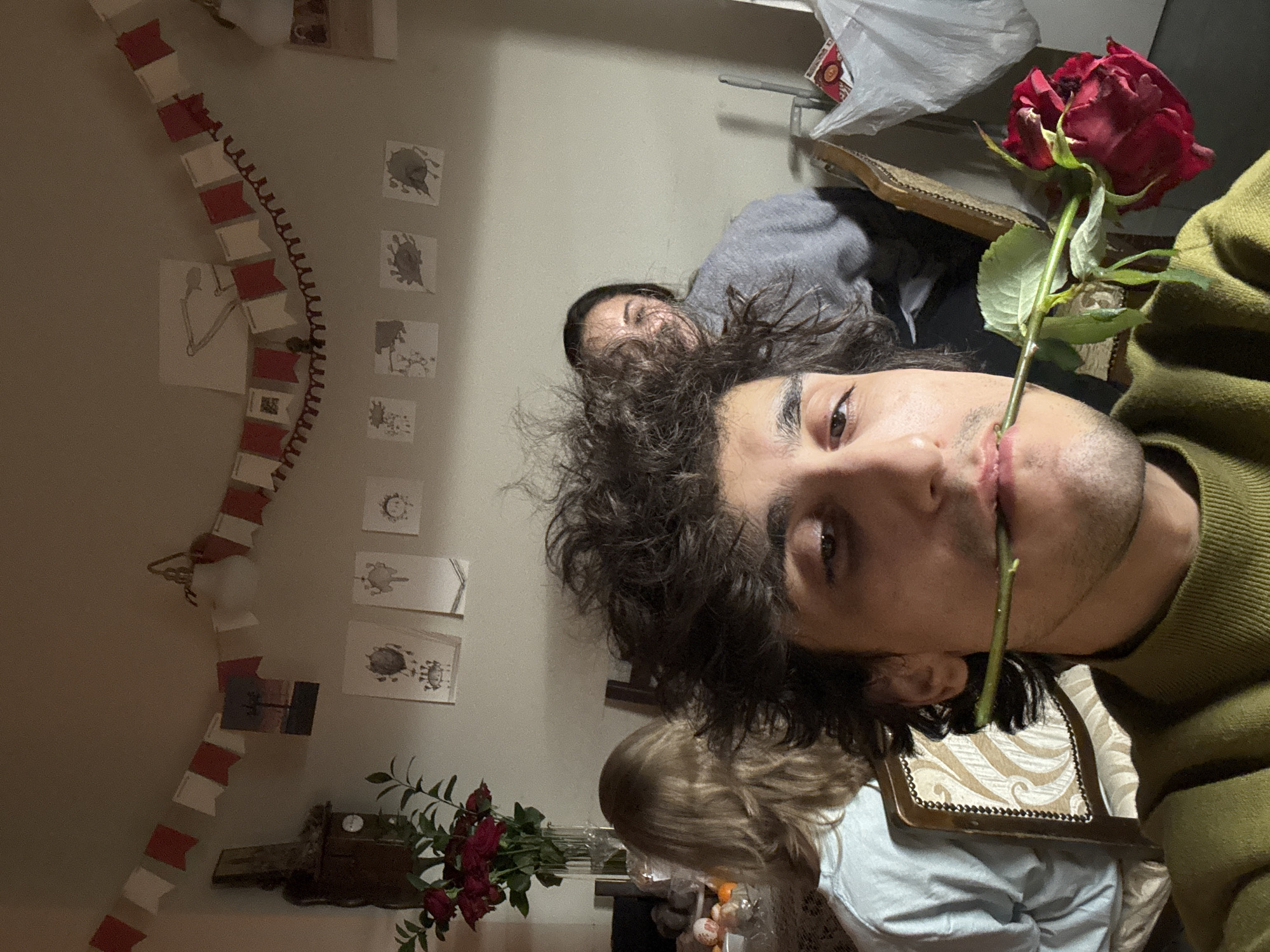 A medium-shot, medium-angle photograph shows a person holding a red rose in their mouth. The person has dark curly hair, brown eyes, and is wearing a green sweater. A second person is present in the background, wearing a grey top. Behind the person in the background, there is what appears to be a white grocery bag on a metal rack. Below, another person is wearing a white shirt and sitting in a decorated wooden chair. In the left background, there are a row of drawings and paintings hanging on the wall, along with a small bouquet of red roses. There is no indication that this photo was taken in Moscow.