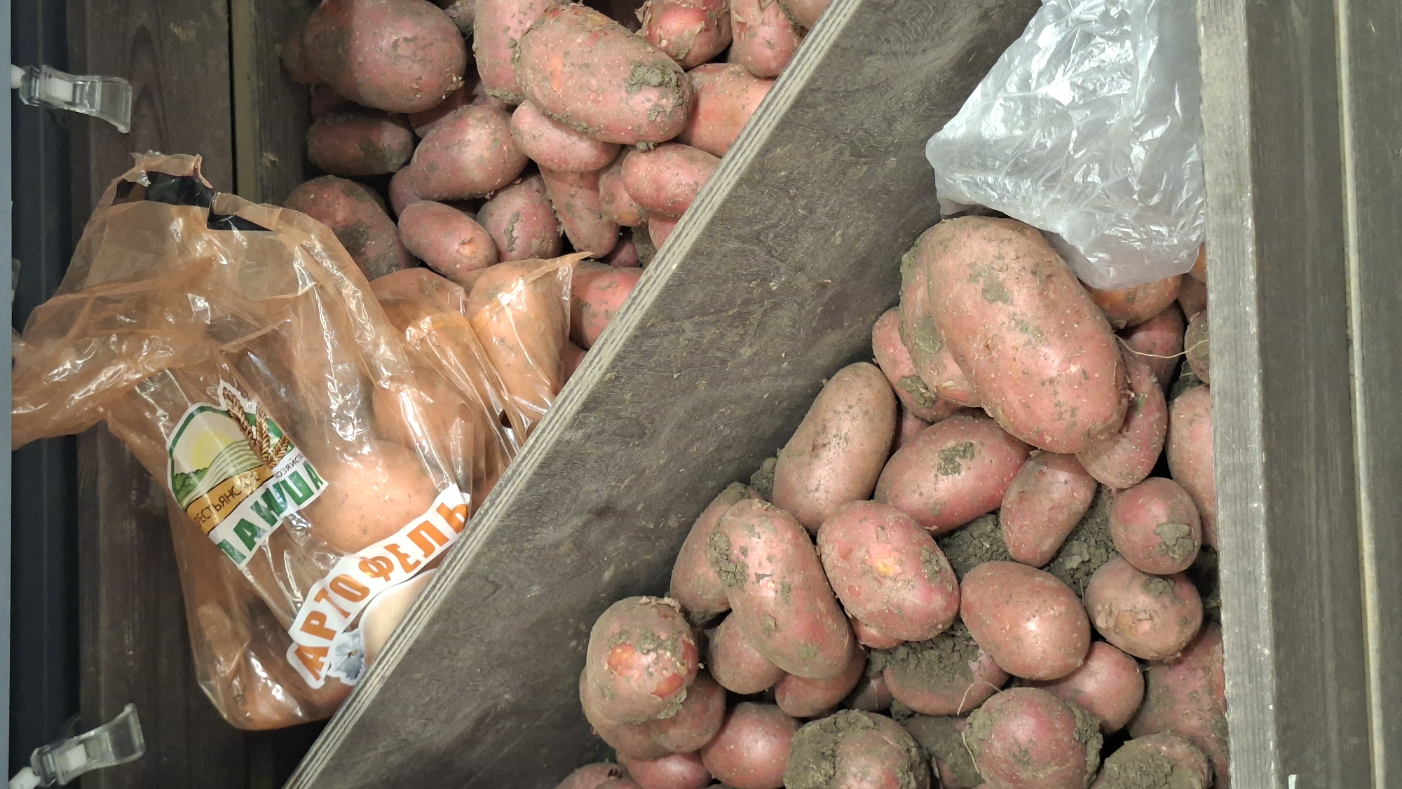 The image shows a close-up view of a wooden crate filled with red potatoes. Some potatoes are still covered in dirt, indicating they are freshly harvested. A plastic bag, also containing potatoes and featuring some Cyrillic text and an image of wheat, is placed on top of some of the potatoes. The crate appears to be part of a market stall or a retail display, possibly in a city like Smalyavichy, Belarus, given the presence of Cyrillic text on the packaging. The lighting suggests an indoor environment.