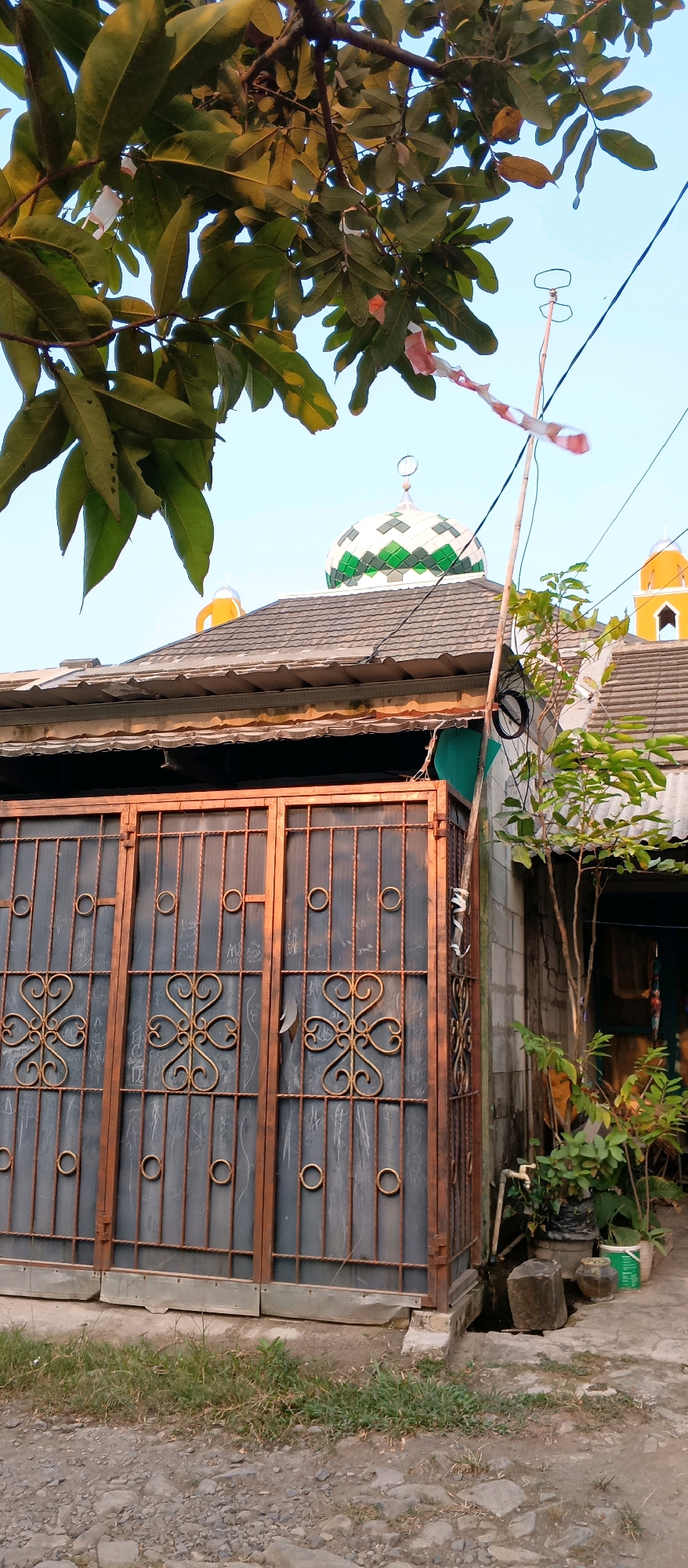 This is a vertical, medium-shot, eye-level photograph of a mosque in Watubelah, Indonesia. The foreground features a section of a tall, metal gate with a rusted copper finish. The gate is composed of vertical bars and decorative elements, including circular rings and ornate floral patterns. To the right of the gate, a small tree with green leaves grows in a pot. Behind the gate and the tree, a cinder block wall is visible. The middle ground is dominated by the roof of a building, with a gray, tiled surface. A mosque dome, featuring a white and green patterned design and a crescent moon on top, rises from behind the roof. A yellow-domed structure is also visible to the right of the mosque dome. In the upper part of the frame, tree branches with green leaves are prominent. The sky is a clear, pale blue. A thin, bamboo pole with a red and white striped ribbon tied to it is positioned to the right of the mosque dome, extending upwards. The overall scene depicts a residential area with a visible religious structure.