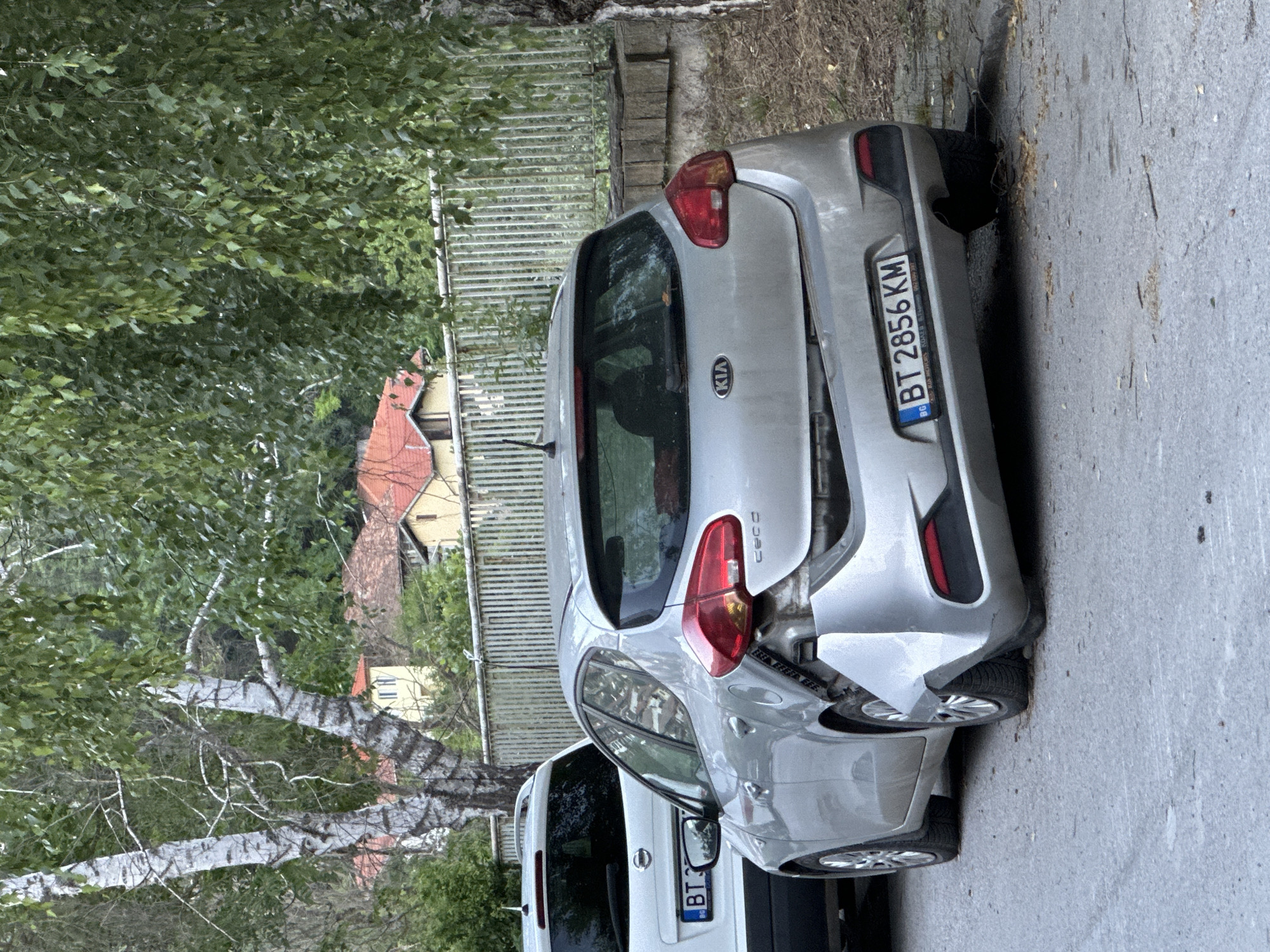 In Veliko Tarnovo, Bulgaria, a silver Kia hatchback is parked on a street, facing right, with significant rear-end damage. The rear bumper is detached and hanging, and the tailgate is visibly bent and warped. A white SUV with a license plate starting with "BT" is parked to the left of the Kia. The scene is lined with trees and a metal fence, with buildings visible in the background. The ground is asphalt, with scattered leaves and debris. The image appears to be taken in daylight.