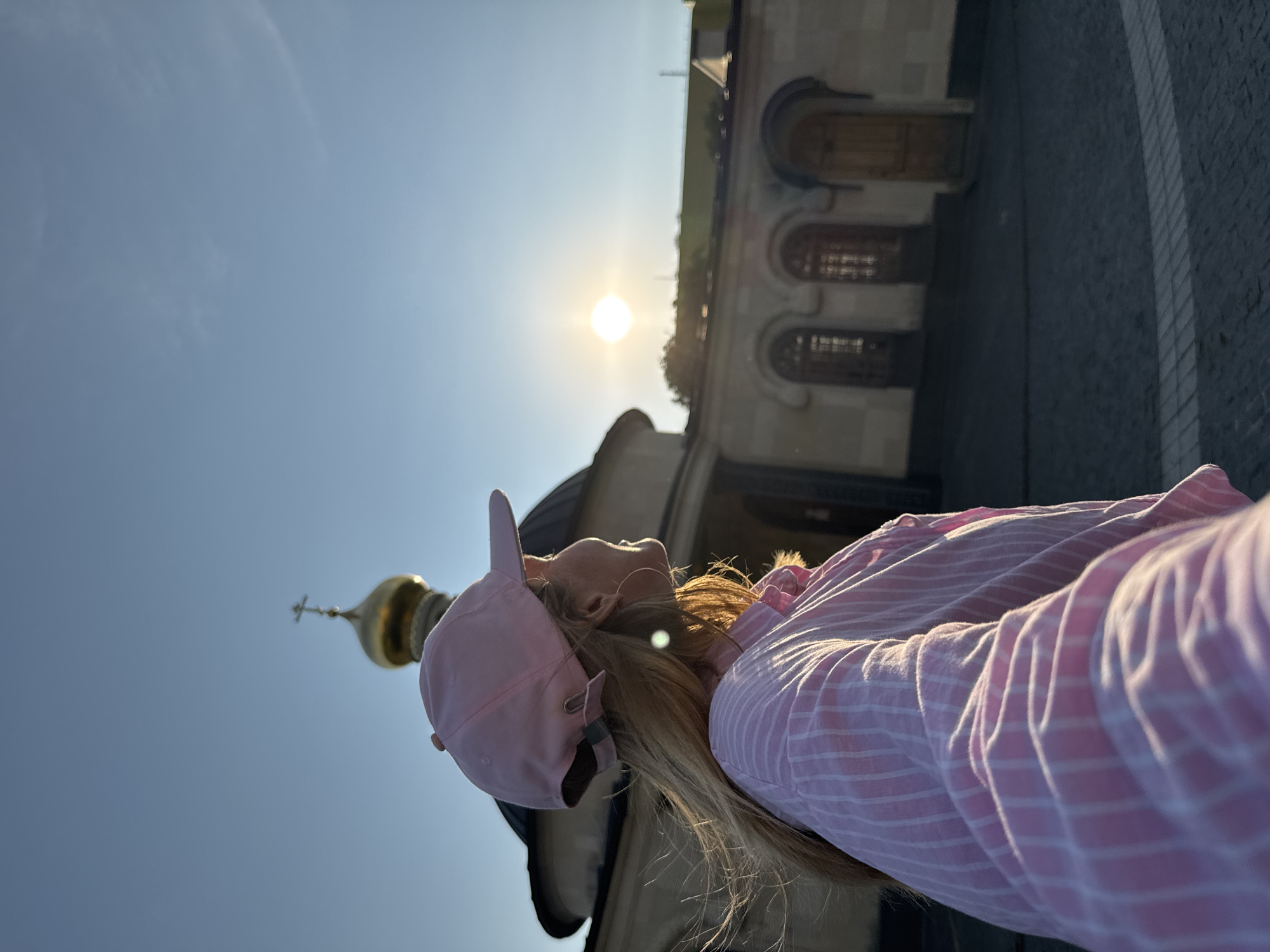 The image shows a woman wearing a pink baseball cap and a pink and white striped shirt, standing in front of a building with a golden dome and cross, likely a church or monastery, in Kirishi, Russia. The sun is low in the sky, casting a warm light on the scene. The woman is looking towards the sun, and her hair is blowing in the wind. The building appears to be made of stone, with arched windows and wooden doors. The ground in front of the building is paved with cobblestones. The overall atmosphere is serene and peaceful.