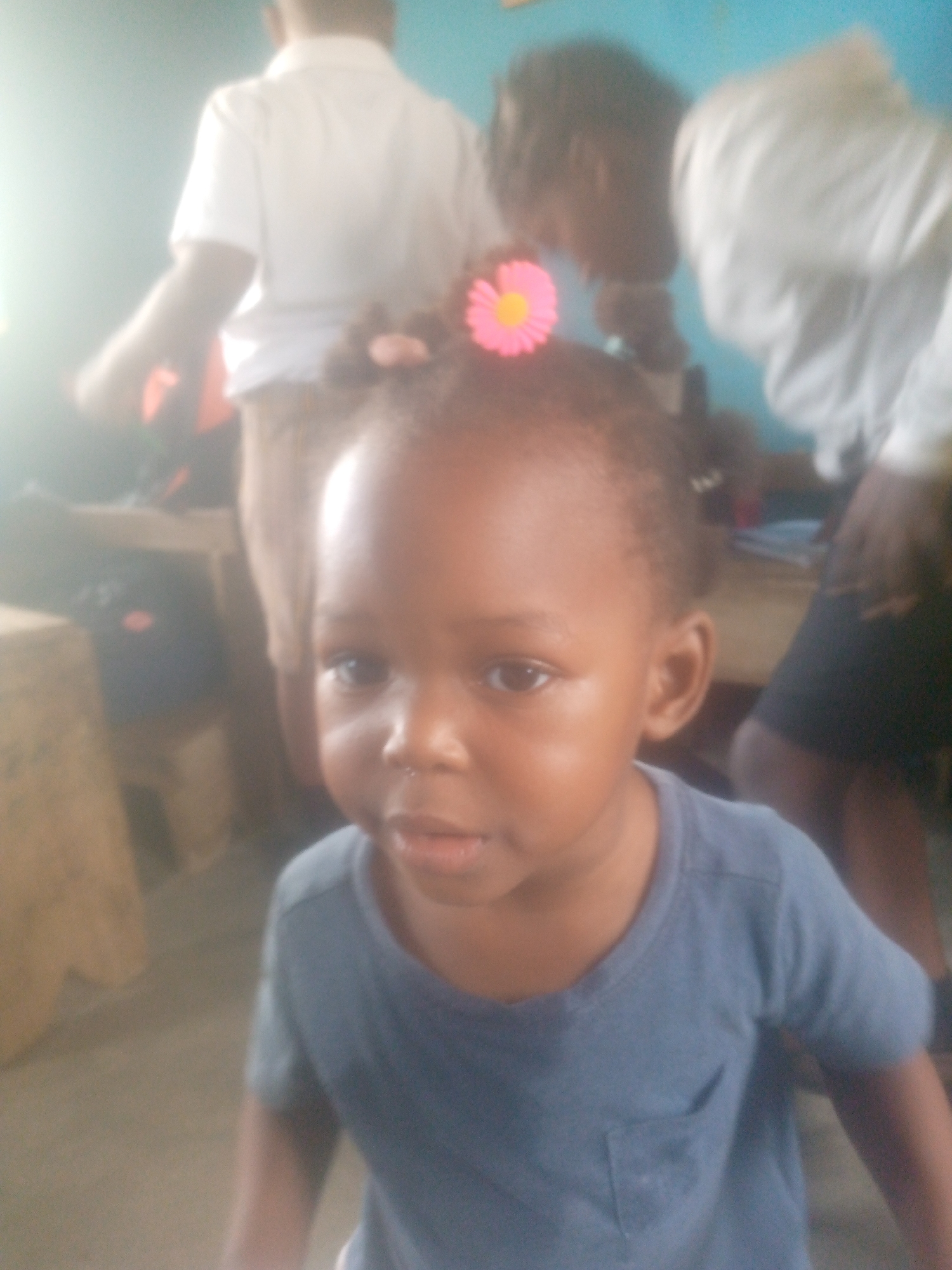 A close-up, slightly blurry image features a young child, likely a toddler, looking directly at the camera in an indoor setting in Ikorodu, Nigeria. The child has dark skin and is wearing a faded blue short-sleeved t-shirt with a small pocket on the left chest. Their hair is styled in small braids or twists, adorned with a vibrant pink and yellow flower hair tie or clip positioned on top of their head. Their expression is neutral and observant.
The background is out of focus but reveals other individuals and furniture. To the left, a blurred figure of a person wearing a white short-sleeved shirt and light-colored shorts or pants is visible from the back. Another indistinct figure, also in a white top, is present on the right side. Vague shapes of wooden tables or stools are discernible in the soft-focus background. The wall behind the subjects appears to be a light blue or teal color.
A bright glare emanates from the left side of the frame, suggesting a strong light source such as a window, contributing to the diffused lighting of the scene. The image captures a candid moment in what appears to be a casual group setting. There is no visible text.