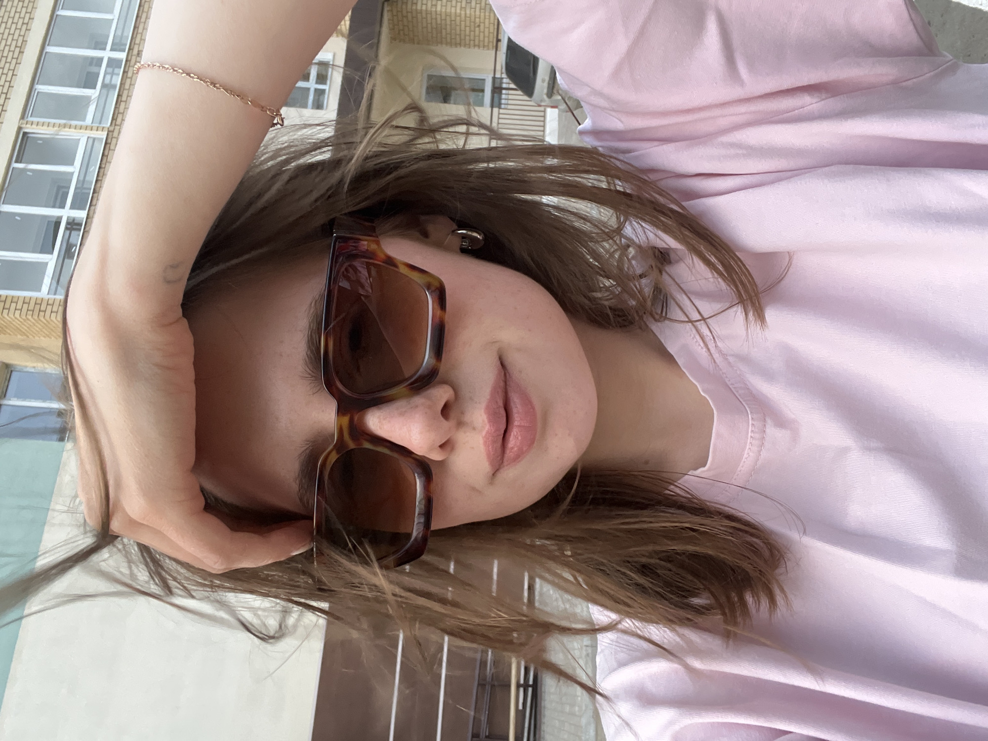 A woman with brown hair and sunglasses is posing for a selfie outdoors. She is wearing a light pink t-shirt and a thin, gold bracelet. Her right hand is near her forehead. In the background, there is a building with many windows. The location is Chaikovsky, Russia.